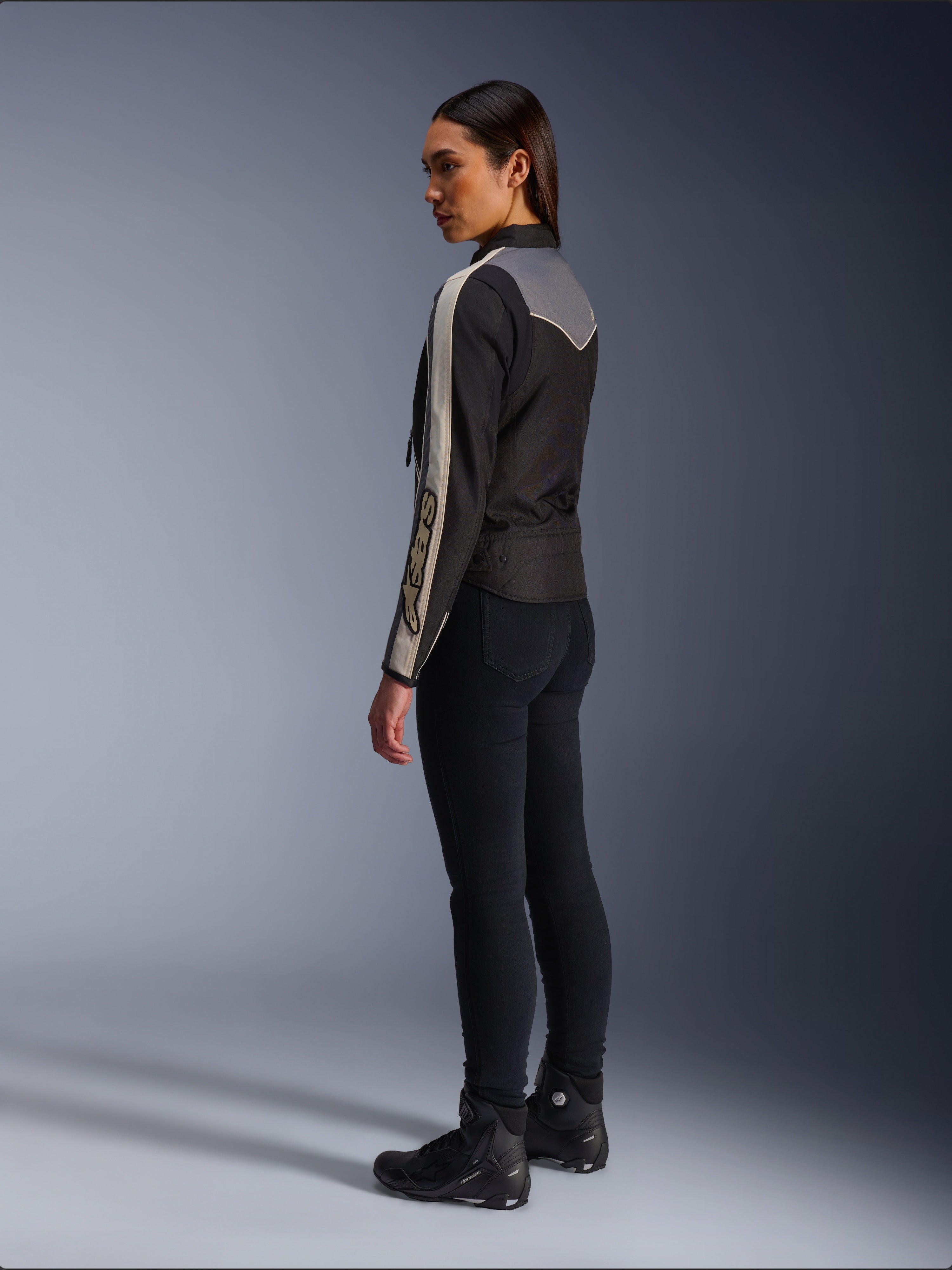 Mannequin femme vue de profil et de dos portant la veste Alpinestars Stella T-Dyno WR en Noir Gris Foncé Écru, au design d'inspiration vintage avec des bandes écrues sur les manches et un empiècement gris dans le dos, portée avec un pantalon noir et des chaussures de moto noires.