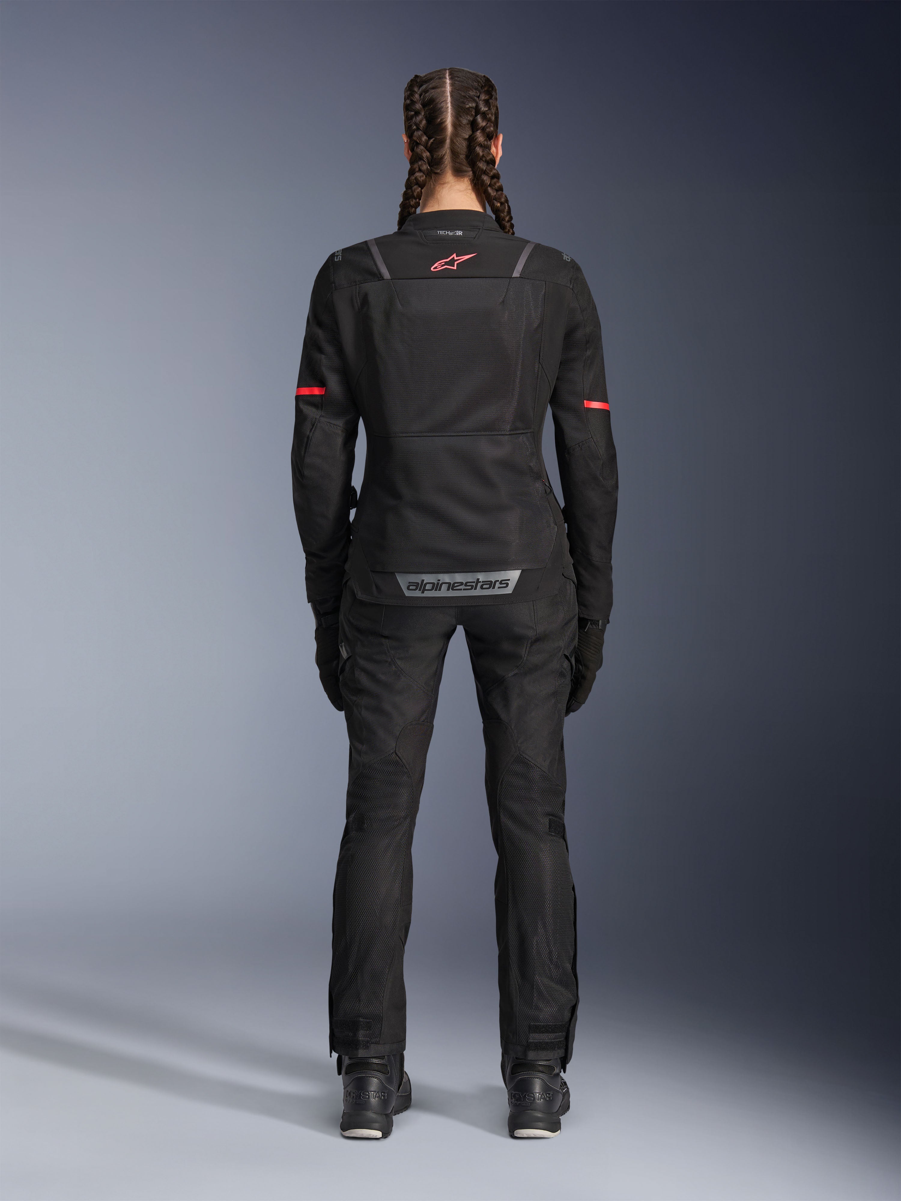 Veste Alpinestars Stella ST-2 Air, veste de moto pour femme, noire, vue arrière présentant un logo rouge sur le haut du dos et un marquage gris sur le bas du dos, portée avec un pantalon et des bottes de moto noirs assortis, design touring moderne avec panneaux en mesh et détails réfléchissants