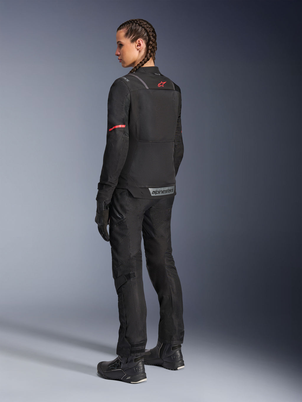 Femme pilote portant un pantalon Alpinestars Stella Andes Air Drystar® noir et une veste Stella ST-2 Air assortie, vue arrière montrant la construction textile technique et les panneaux en maille, logo étoile rouge au dos de la veste, avec bottes et gants de moto noirs.