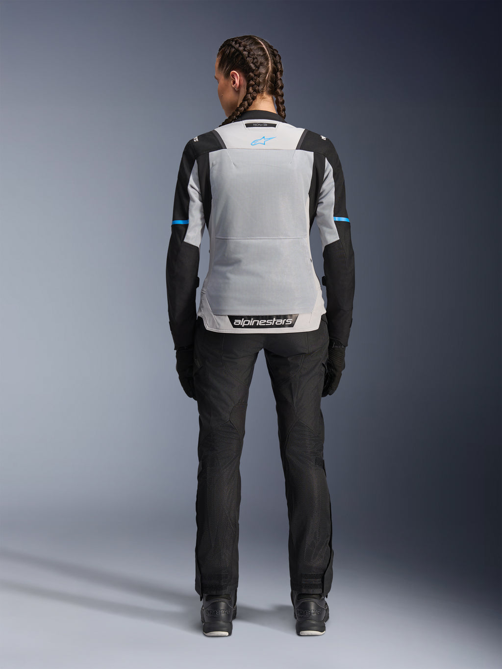 Veste Alpinestars Stella ST-2 Air, Ice Gray Black, vue arrière sur modèle féminin, avec un large panneau arrière en mesh gris clair, manches noires avec des bandes bleues, marquage Alpinestars sur le bas du dos, associée à un pantalon technique et des bottes de moto noirs, équipement de touring moto d'été
