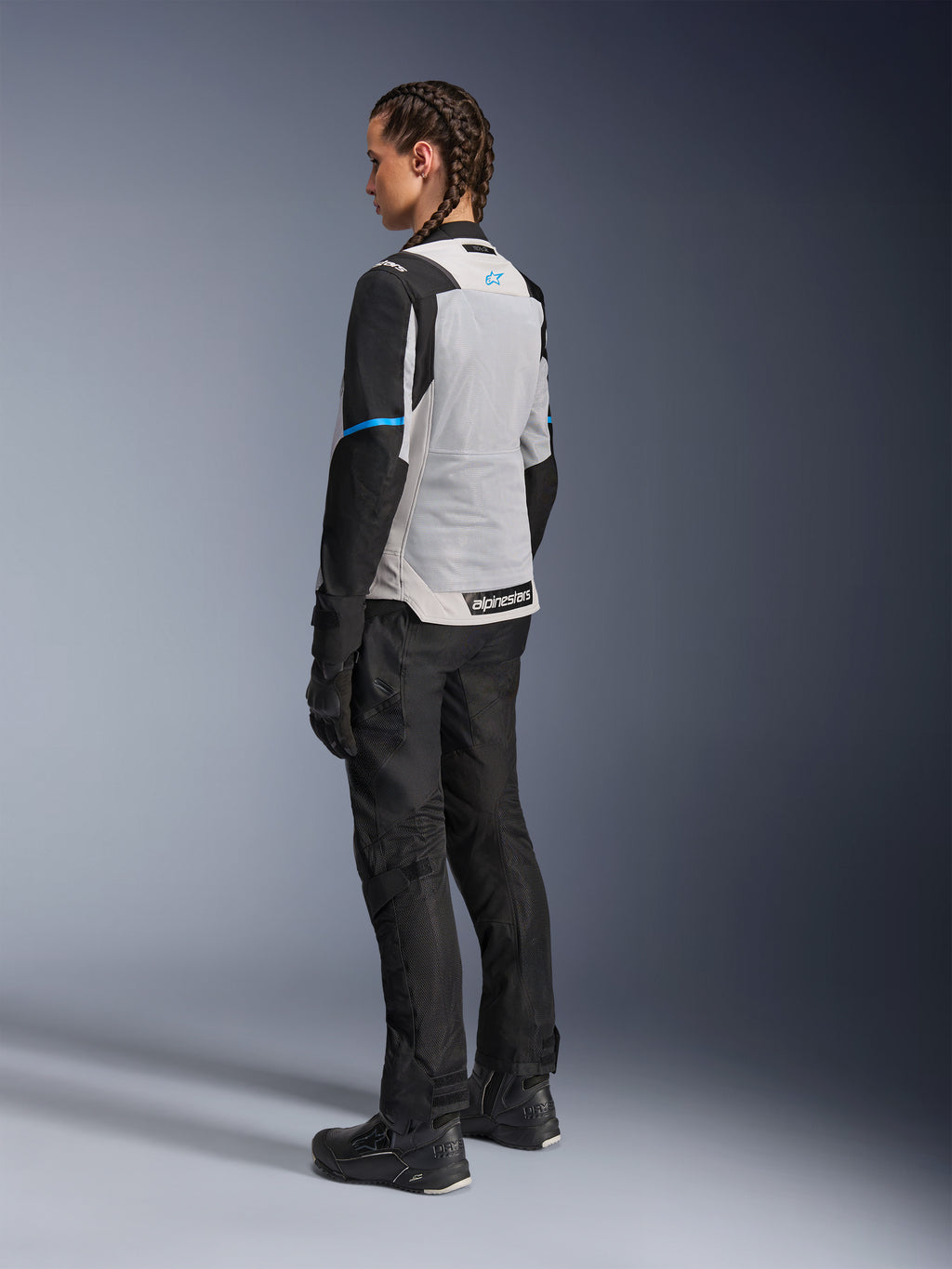 Motarde portant la veste Alpinestars Stella ST-2 Air en Gris Glace et Noir, vue de dos de trois-quarts mettant en valeur le large panneau arrière en maille gris clair, les manches noires avec une bande bleue et le logo Alpinestars bleu, associée à un pantalon, des gants et des bottes de moto noirs pour une tenue d'aventure touring complète.