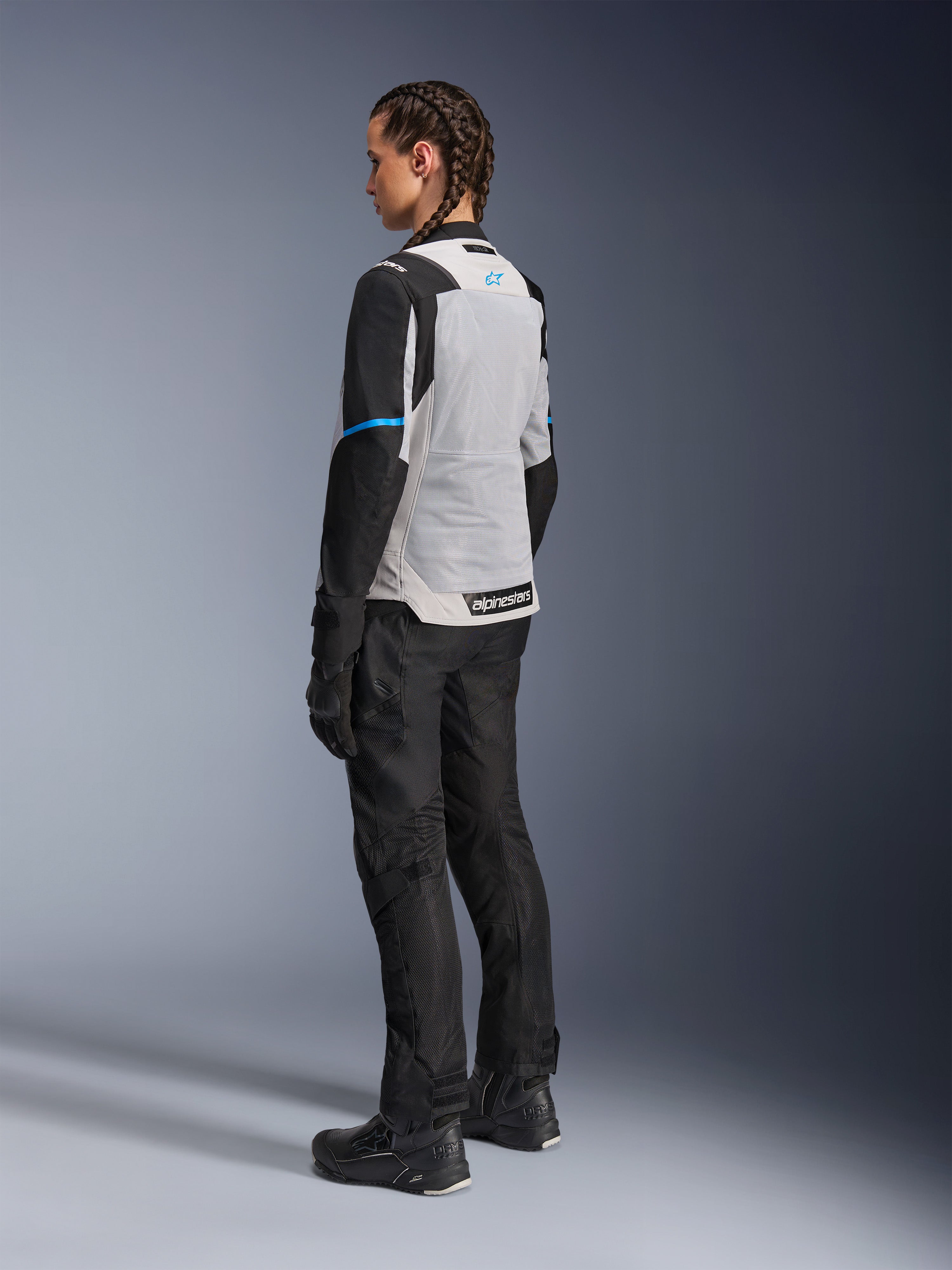 Motarde portant la veste Alpinestars Stella ST-2 Air en Gris Glace et Noir, vue de dos de trois-quarts mettant en valeur le large panneau arrière en maille gris clair, les manches noires avec une bande bleue et le logo Alpinestars bleu, associée à un pantalon, des gants et des bottes de moto noirs pour une tenue d'aventure touring complète.