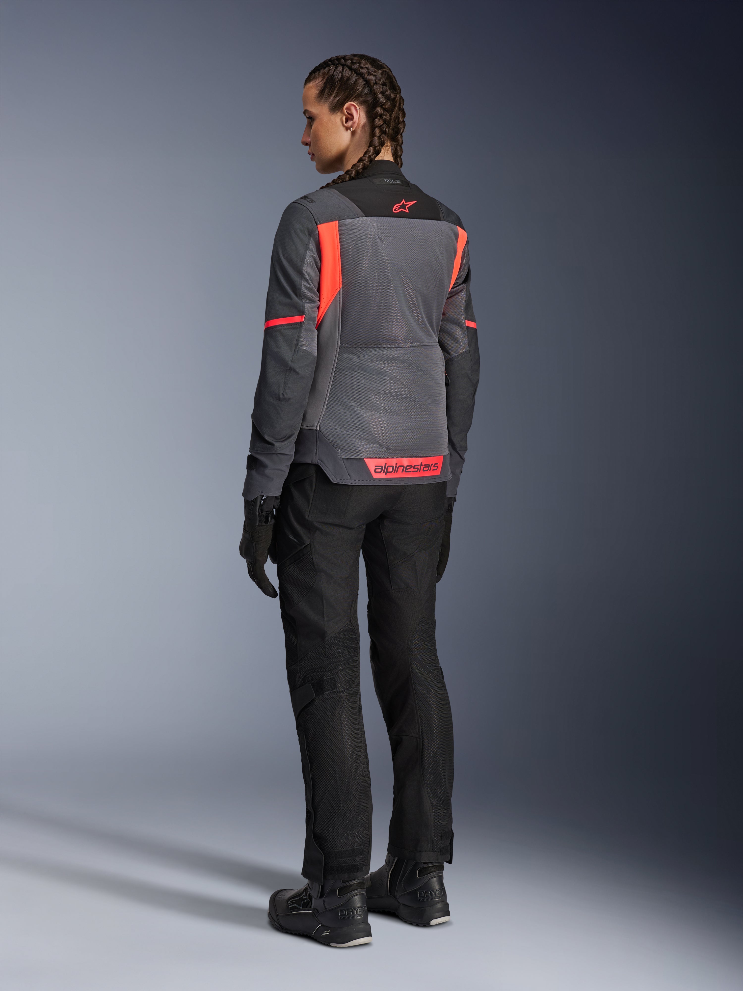 Femme portant la veste Alpinestars Stella St-2 Air, veste de moto touring, gris profond et gris foncé avec des accents corail fluo, associée à un pantalon et des gants de moto noirs, vue arrière montrant de grands panneaux en mesh et le marquage corail réfléchissant, conçue pour les aventures à moto en été