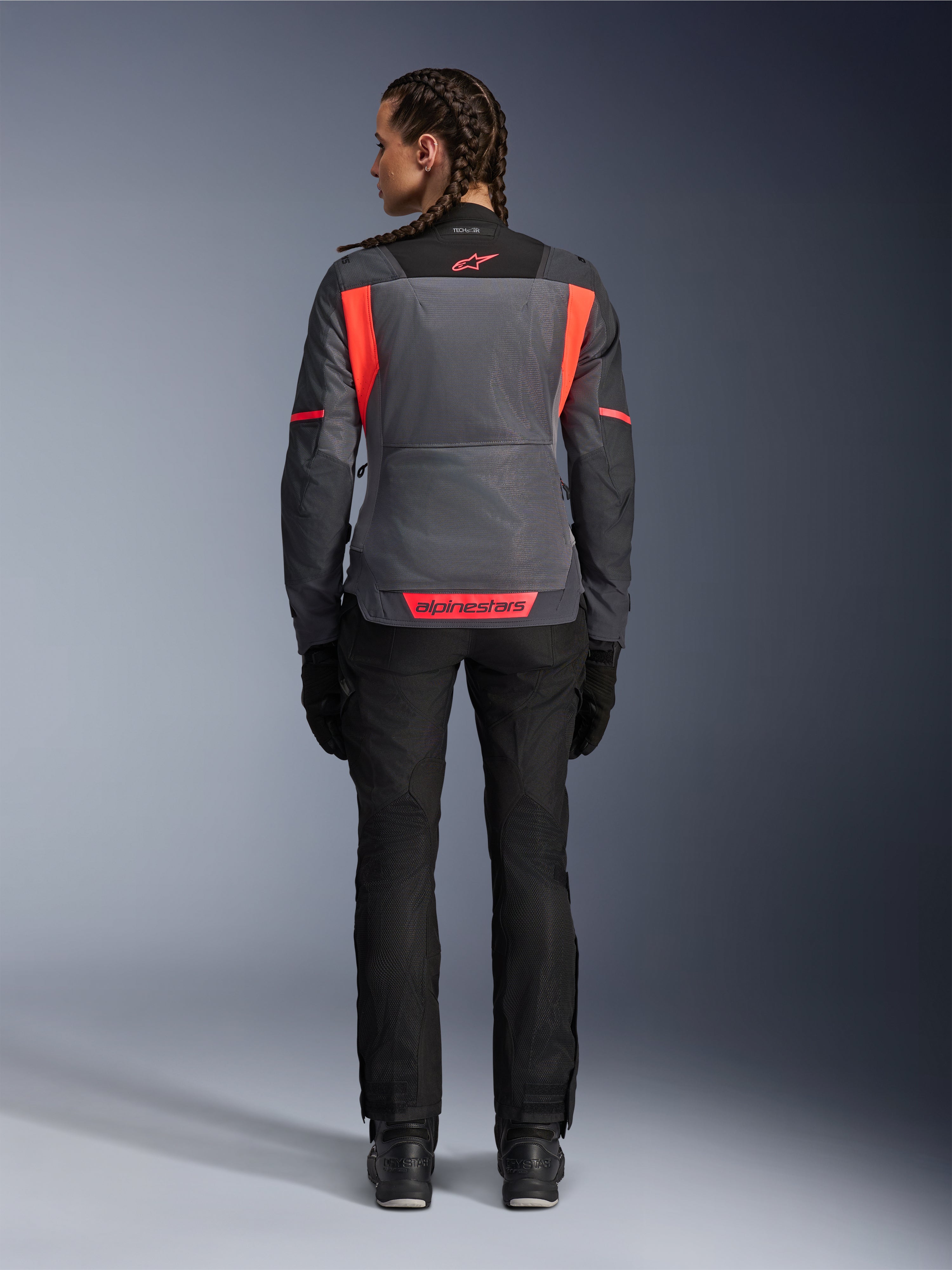 Vue arrière d'un modèle portant la veste Alpinestars Stella St-2 Air pour femme en Gris Profond et Gris Foncé avec des accents Corail Fluo, dotée de larges panneaux de ventilation en mesh dans le dos et de détails de logo réfléchissants, associée à un pantalon et des gants de moto noirs.