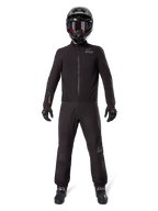 Pilote de moto portant la doublure de pantalon Alpinestars AMT-8 Stretch Drystar® XF et la veste assortie, équipement technique noir imperméable et coupe-vent, avec casque, gants et bottes noirs, design adventure touring épuré.