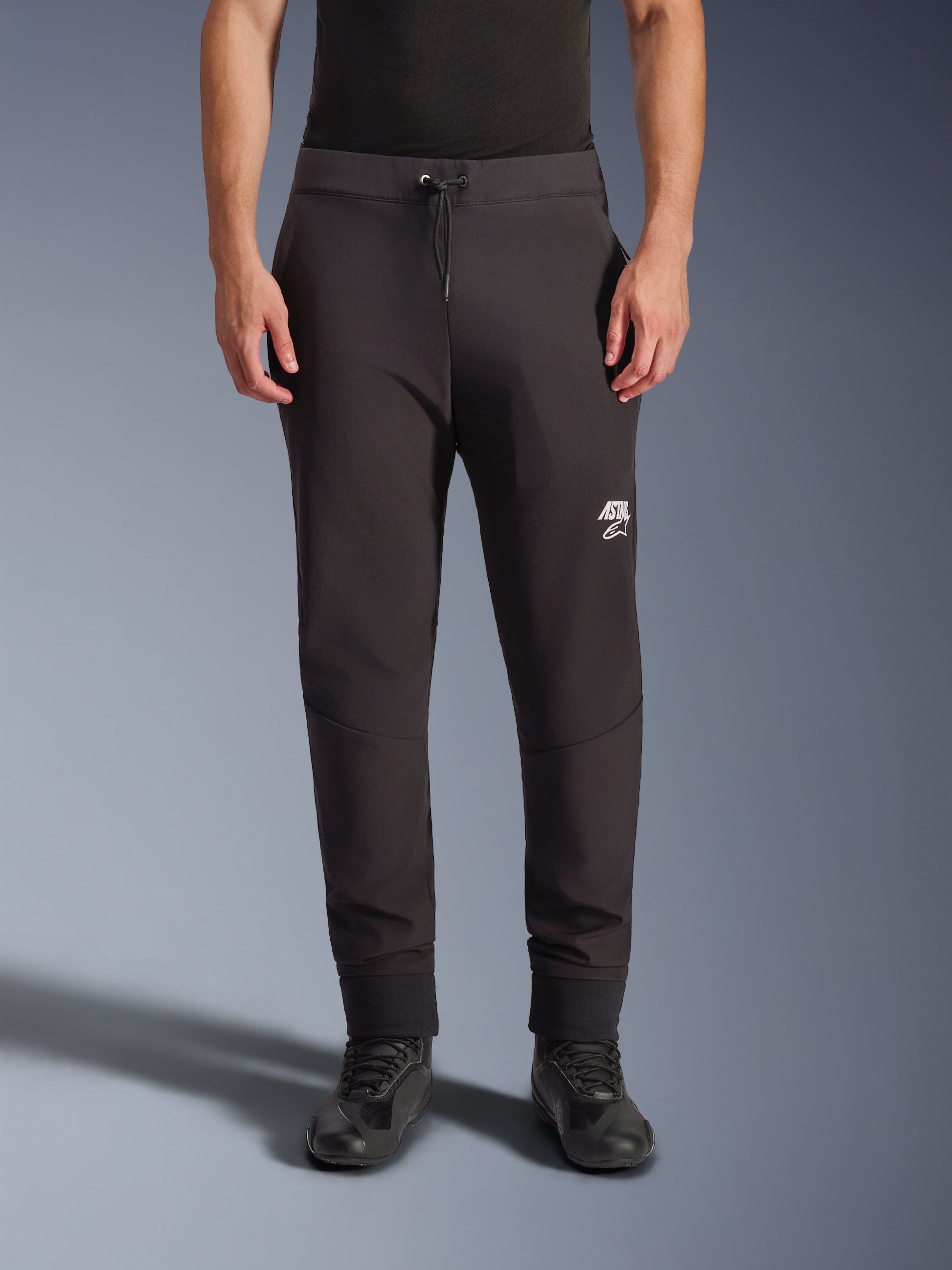 Alpinestars Aeroshell Pants, pantalon de moto en softshell noir, coupe athlétique style jogging avec cordon de serrage et ourlets élastiques, logo Astars blanc, porté avec des chaussures de moto noires, équipement crossover moderne pour la route et un usage décontracté
