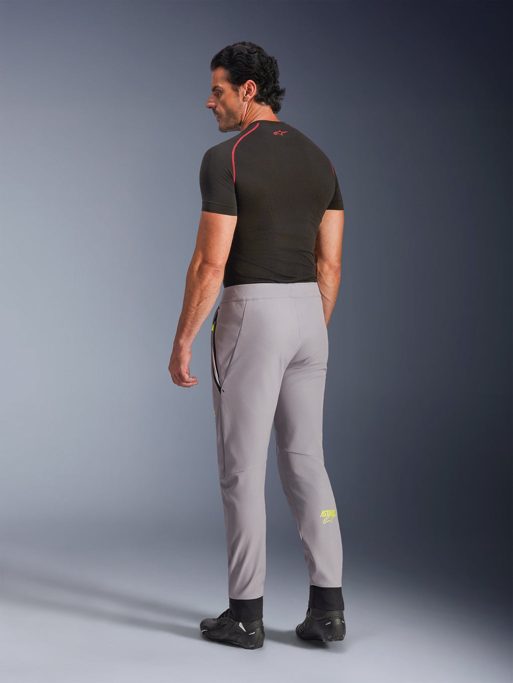 Pantalon Alpinestars Aeroshell, Mid Gray Yellow Fluo, vue de dos d'un mannequin homme portant un pantalon de moto en softshell extensible, avec le logo Astars jaune sur le mollet gauche, associé à une couche de base noire et des chaussures de moto pour une silhouette sportive moderne.