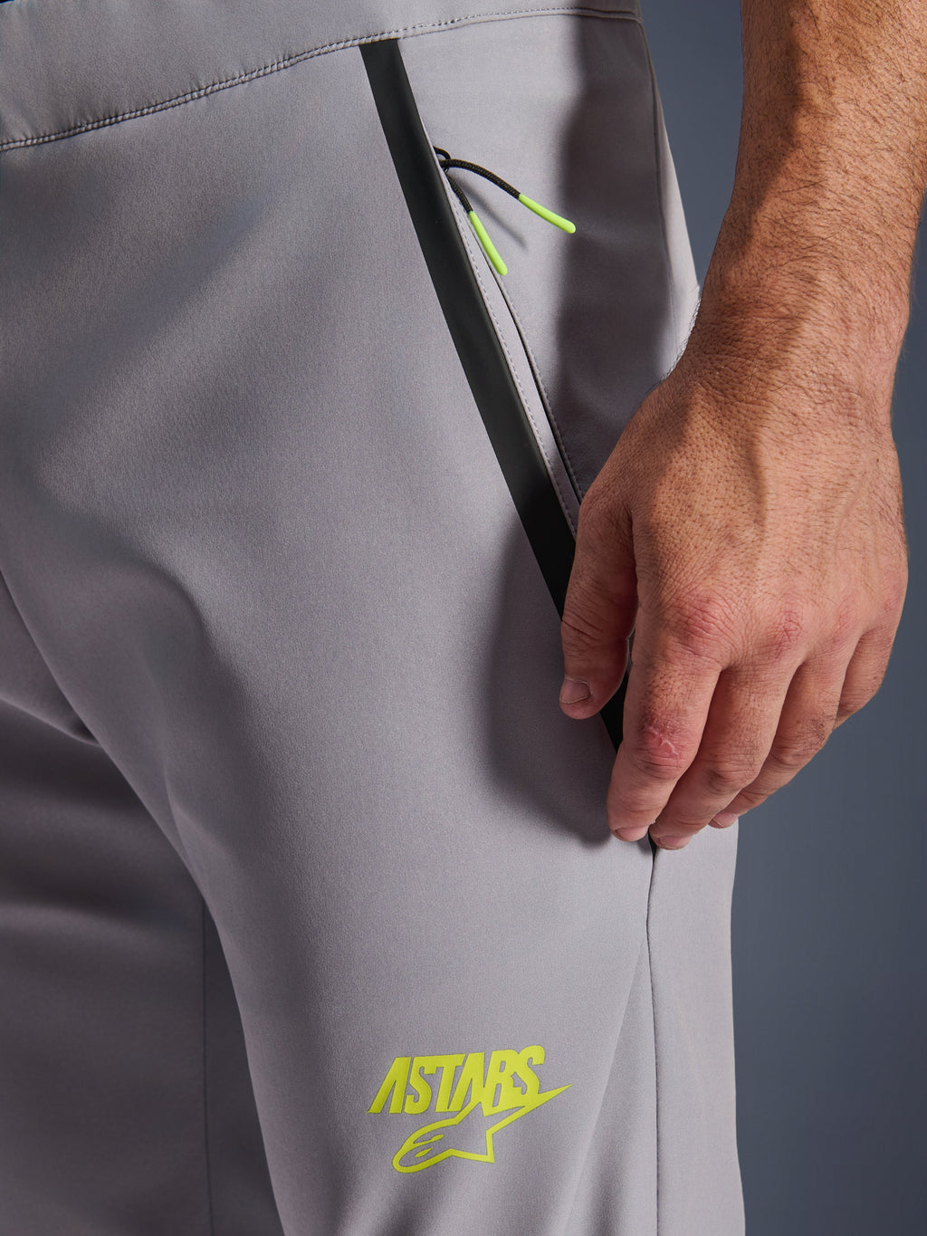 Pantalon Alpinestars Aeroshell, Gris Moyen avec accents Jaune Fluo, vue rapprochée de la poche avant zippée avec bordure noire et cordons jaune vif, logo ASTARS jaune sur la cuisse, matériau softshell extensible pour la pratique de la moto.