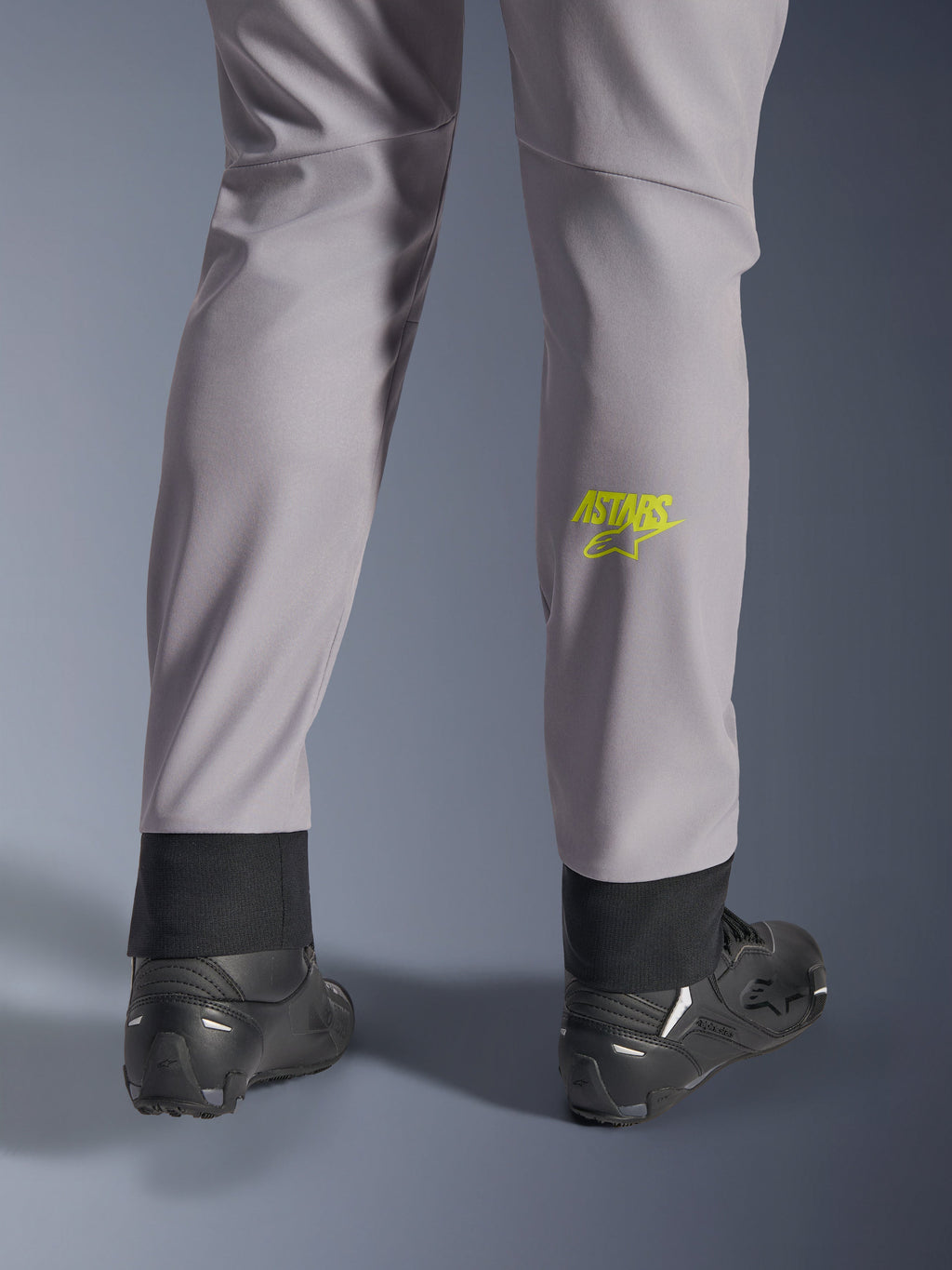 Pantalon Alpinestars Aeroshell, gris moyen avec logo Astars jaune fluo sur le mollet, vue de dos avec chaussures de moto noires, doté d'un ourlet inférieur élastique et d'une silhouette fuselée moderne