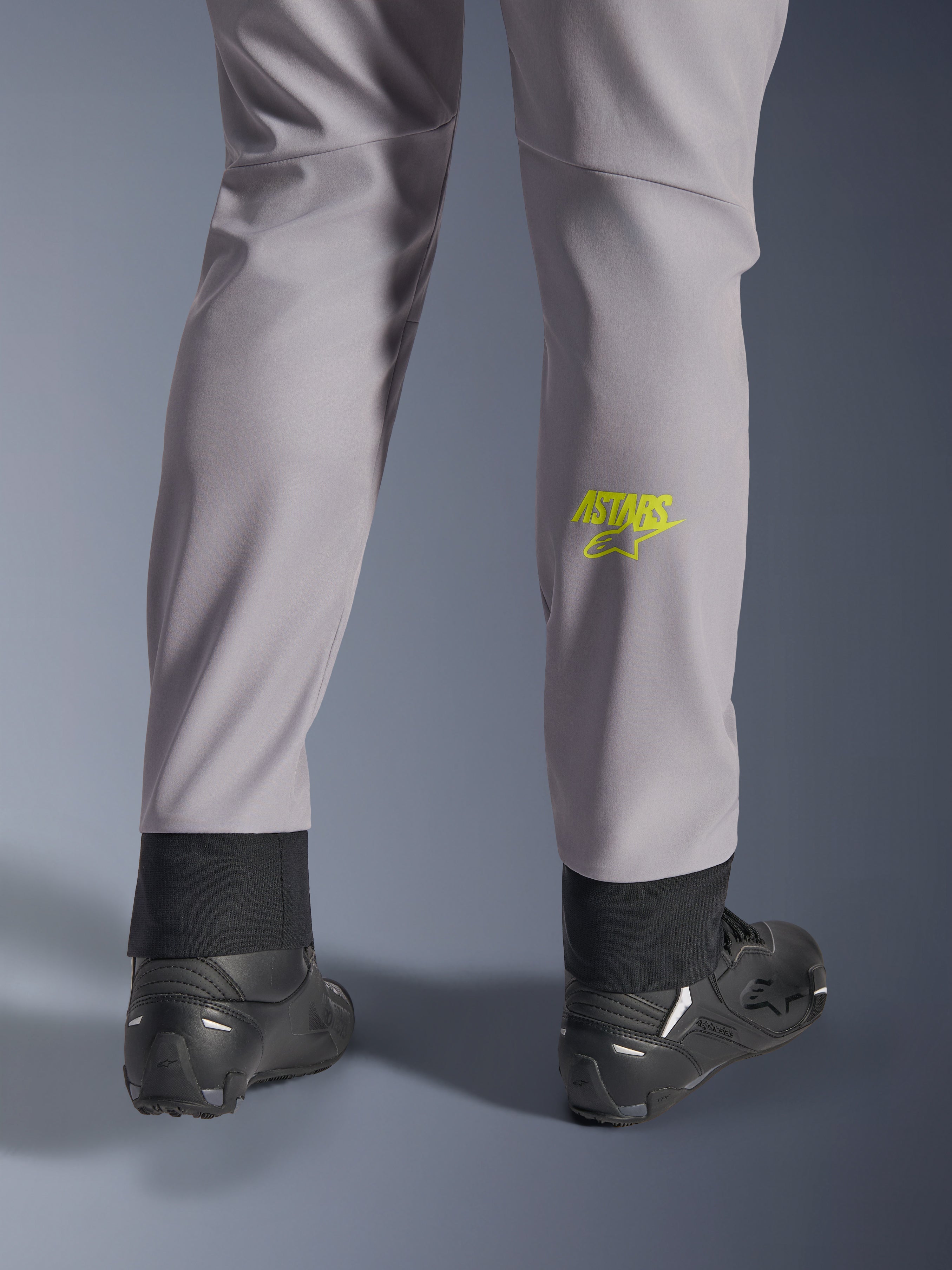 Pantalon Alpinestars Aeroshell, gris moyen avec logo Astars jaune fluo sur le mollet, vue de dos avec chaussures de moto noires, doté d'un ourlet inférieur élastique et d'une silhouette fuselée moderne