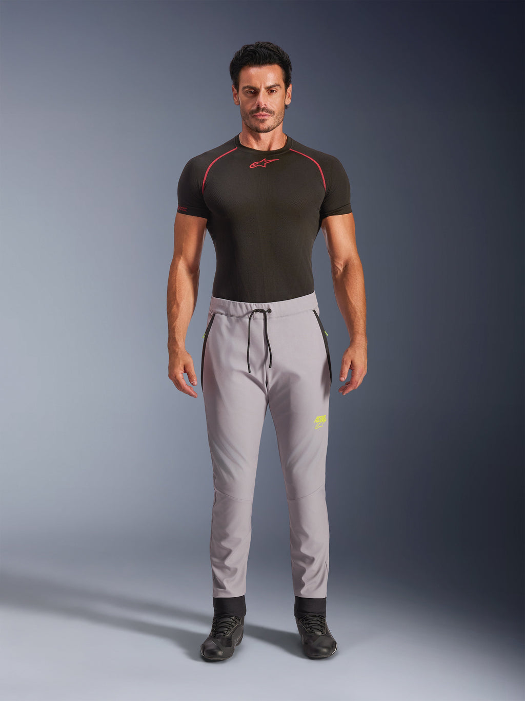 Mannequin homme portant le pantalon Aeroshell Alpinestars en Gris Moyen et Jaune Fluo, pantalon softshell de sport polyvalent avec taille élastique à cordon de serrage et poches zippées, associé à un haut technique noir à manches courtes, design athlétique moderne pour la route et la conduite décontractée
