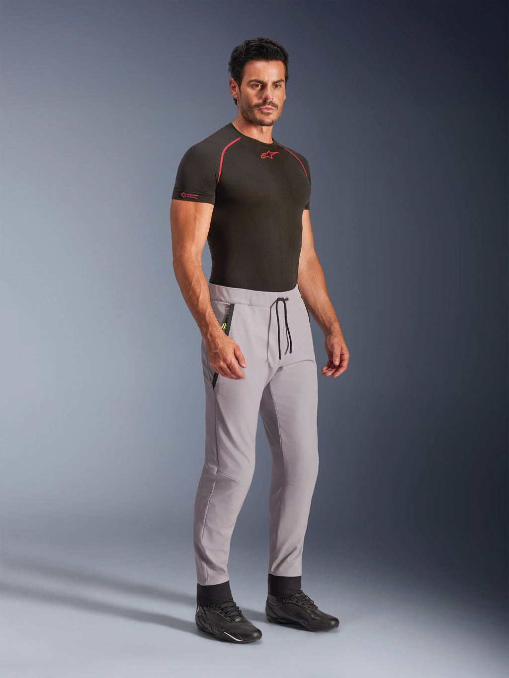 Pantalon Alpinestars Aeroshell, gris moyen avec accents jaune fluo, pantalon softshell polyvalent porté par un mannequin avec une sous-couche noire et des chaussures noires, doté d'une coupe fuselée moderne, d'une taille élastique avec cordon de serrage noir et de poches zippées.