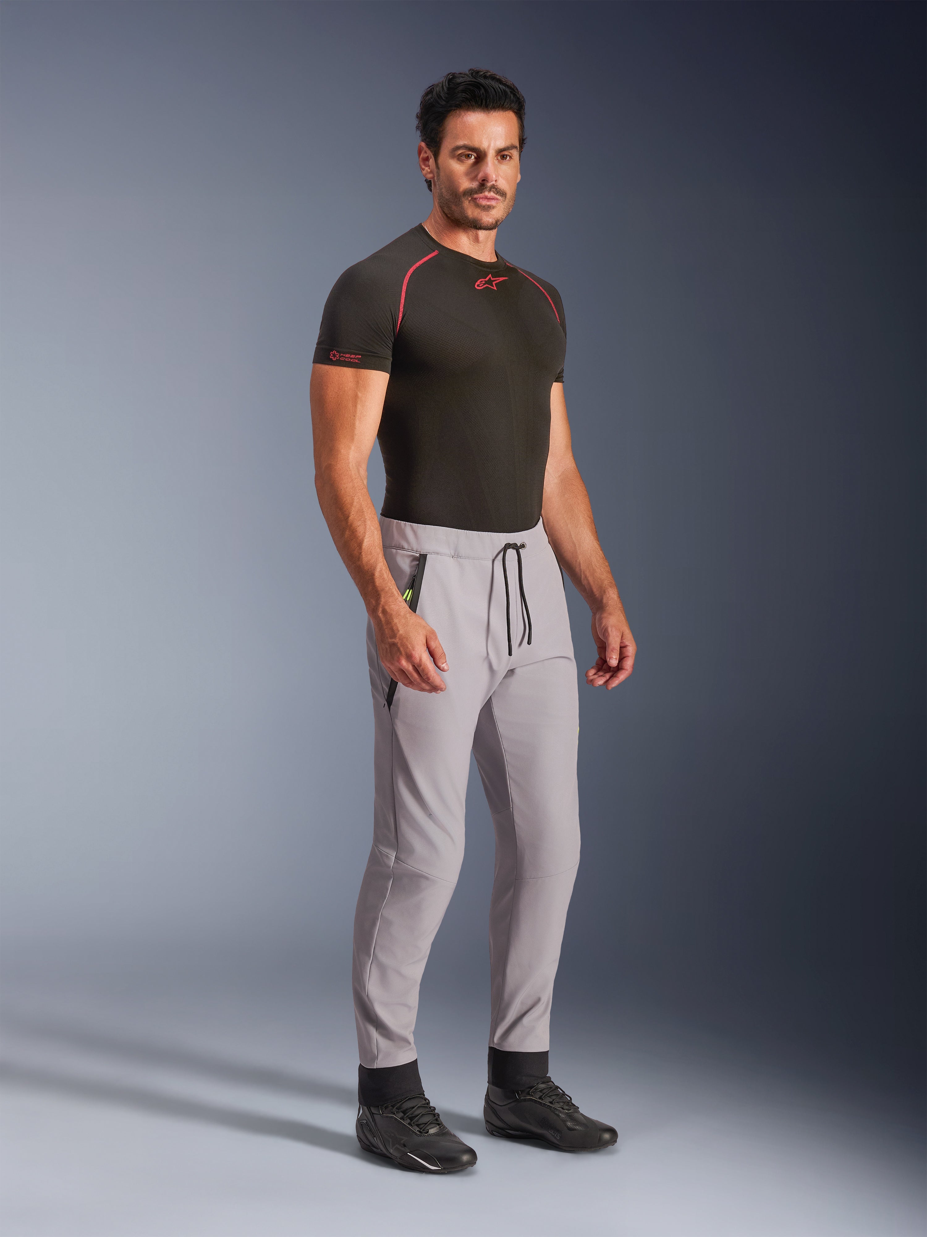 Pantalon Alpinestars Aeroshell, gris moyen avec accents jaune fluo, pantalon softshell polyvalent porté par un mannequin avec une sous-couche noire et des chaussures noires, doté d'une coupe fuselée moderne, d'une taille élastique avec cordon de serrage noir et de poches zippées.