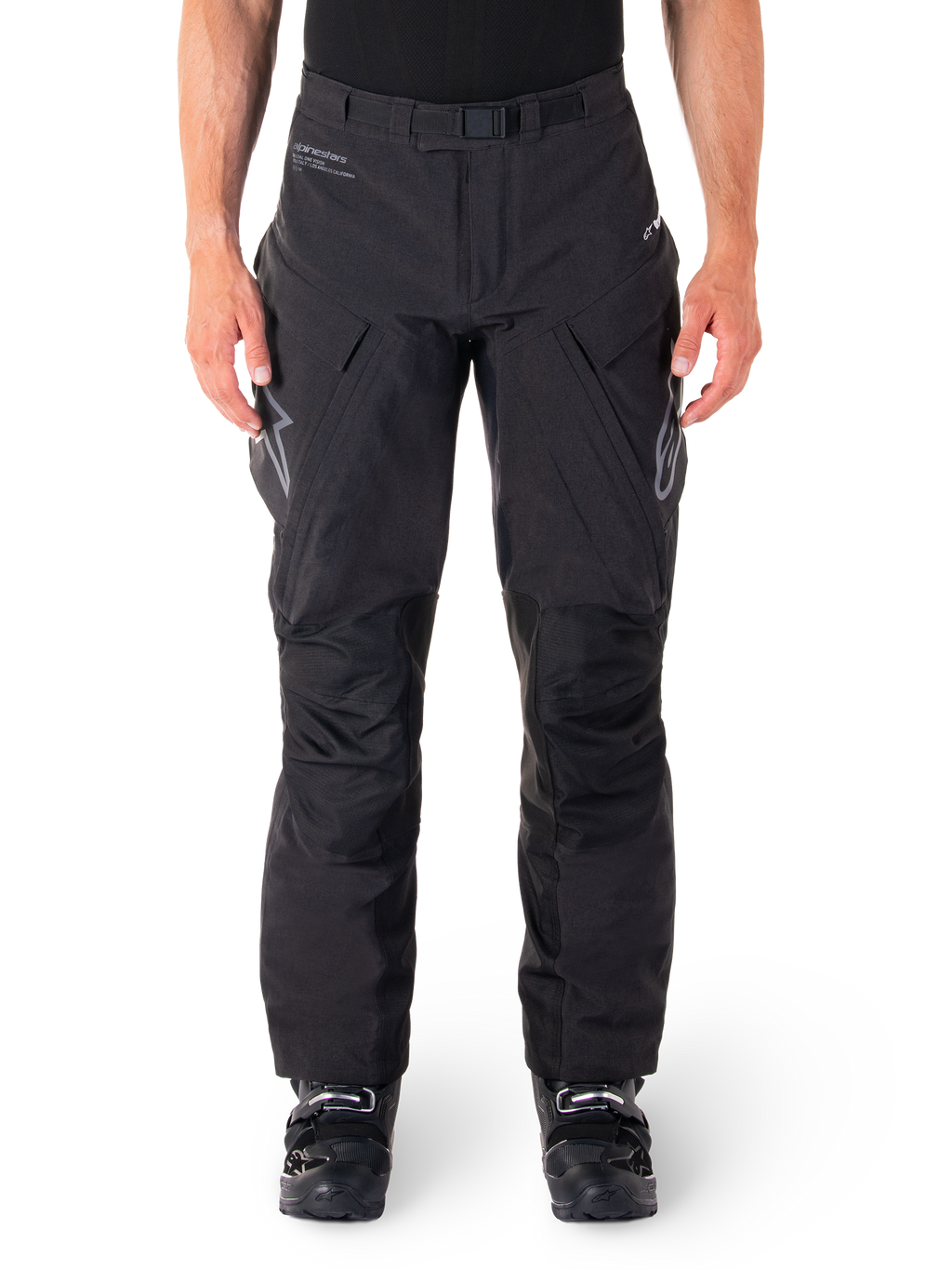 Pantalon Alpinestars Hyde XT Drystar, pantalon d'aventure touring noir, porté par un pilote avec des bottes noires, logos gris sur les cuisses, ceinture intégrée, zones de genoux renforcées et construction textile imperméable.