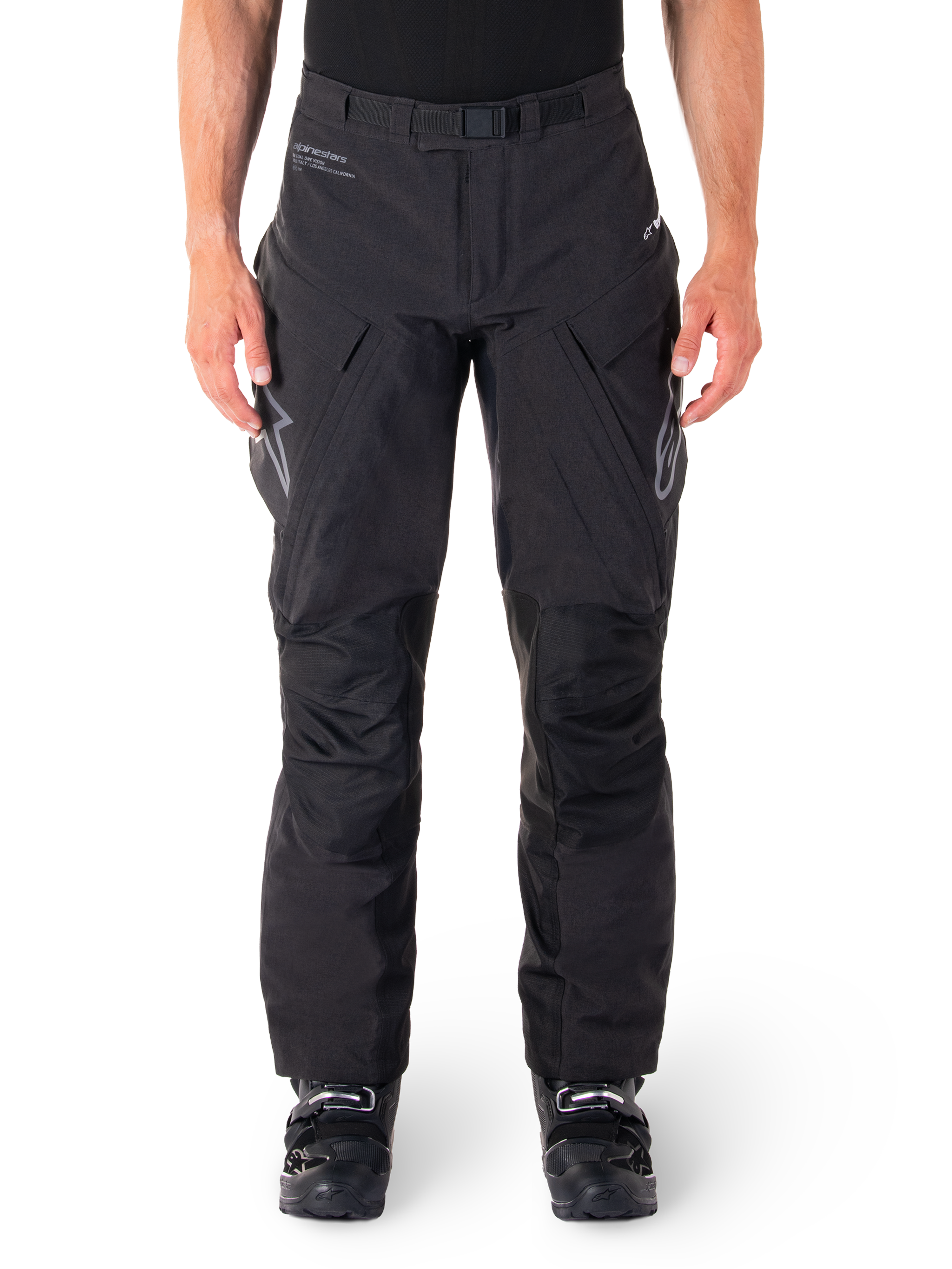 Pantalon Alpinestars Hyde XT Drystar, pantalon d'aventure touring noir, porté par un pilote avec des bottes noires, logos gris sur les cuisses, ceinture intégrée, zones de genoux renforcées et construction textile imperméable.