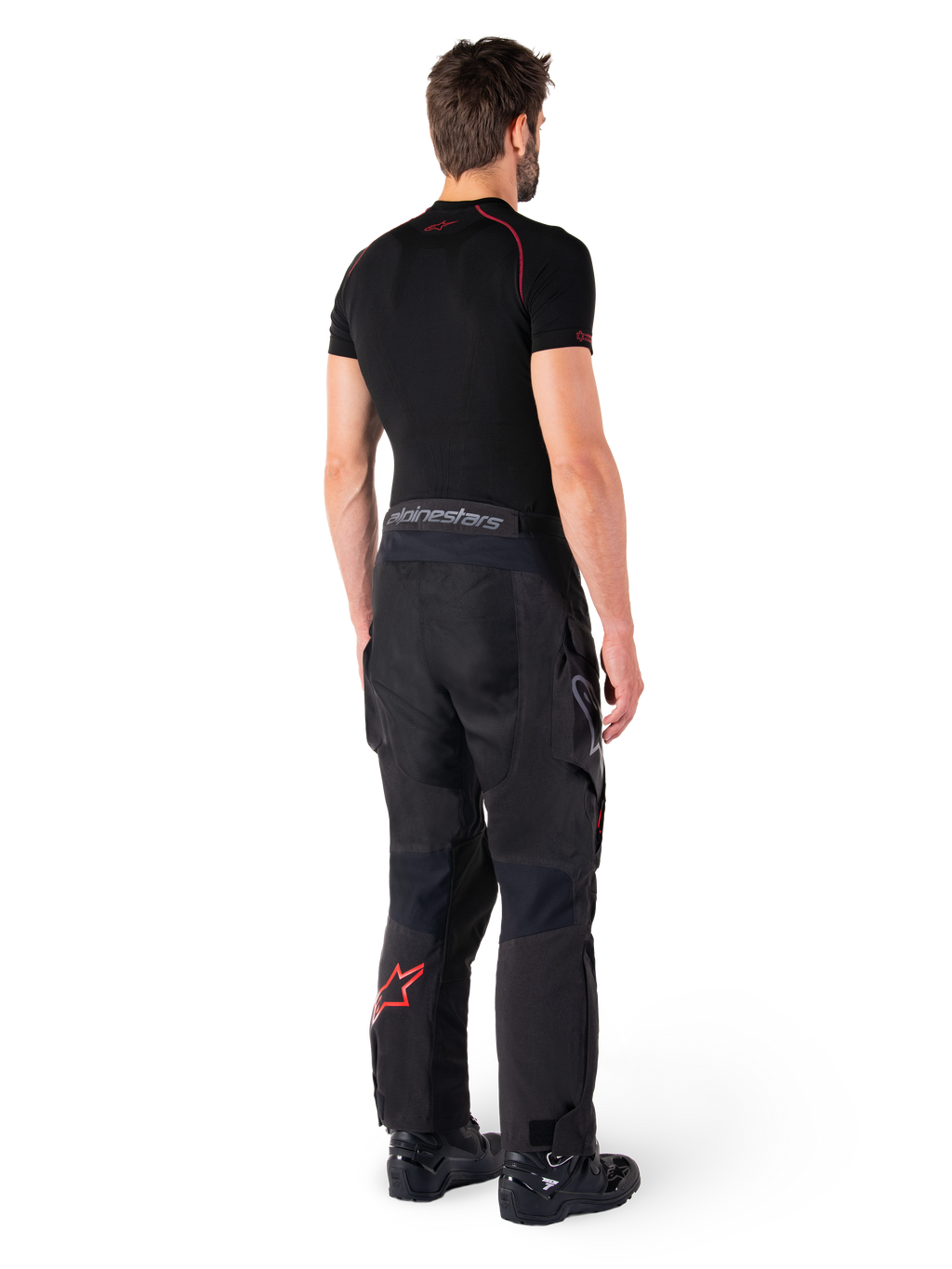 Pantalon Alpinestars Hyde XT Drystar®, pantalon d'aventure touring, noir et rouge vif, vue arrière d'un pilote portant le pantalon avec une sous-couche noire et des bottes noires, doté d'une construction textile durable avec un logo étoile rouge sur le bas de la jambe et le marquage Alpinestars gris sur la ceinture