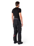 Pantalon Alpinestars Hyde XT Drystar®, pantalon d'aventure touring, noir et rouge vif, vue arrière d'un pilote portant le pantalon avec une sous-couche noire et des bottes noires, doté d'une construction textile durable avec un logo étoile rouge sur le bas de la jambe et le marquage Alpinestars gris sur la ceinture