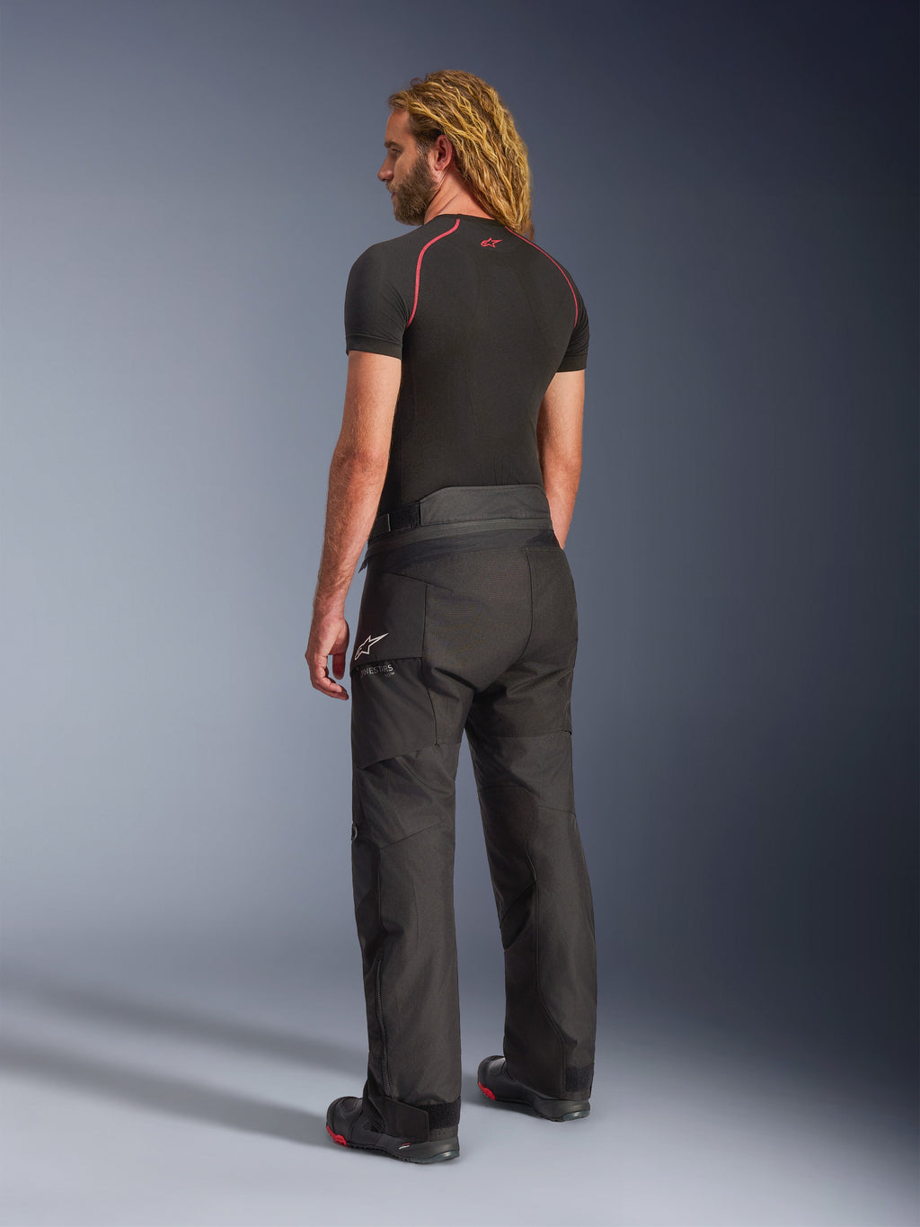 Alpinestars Andes V4 Drystar® Pants, Black motorcycle adventure touring pants, worn by a model from a rear-side view, featuring a white Alpinestars logo on the hip, paired with a black short-sleeve base layer and riding boots, durable textile construction for road and adventure use.