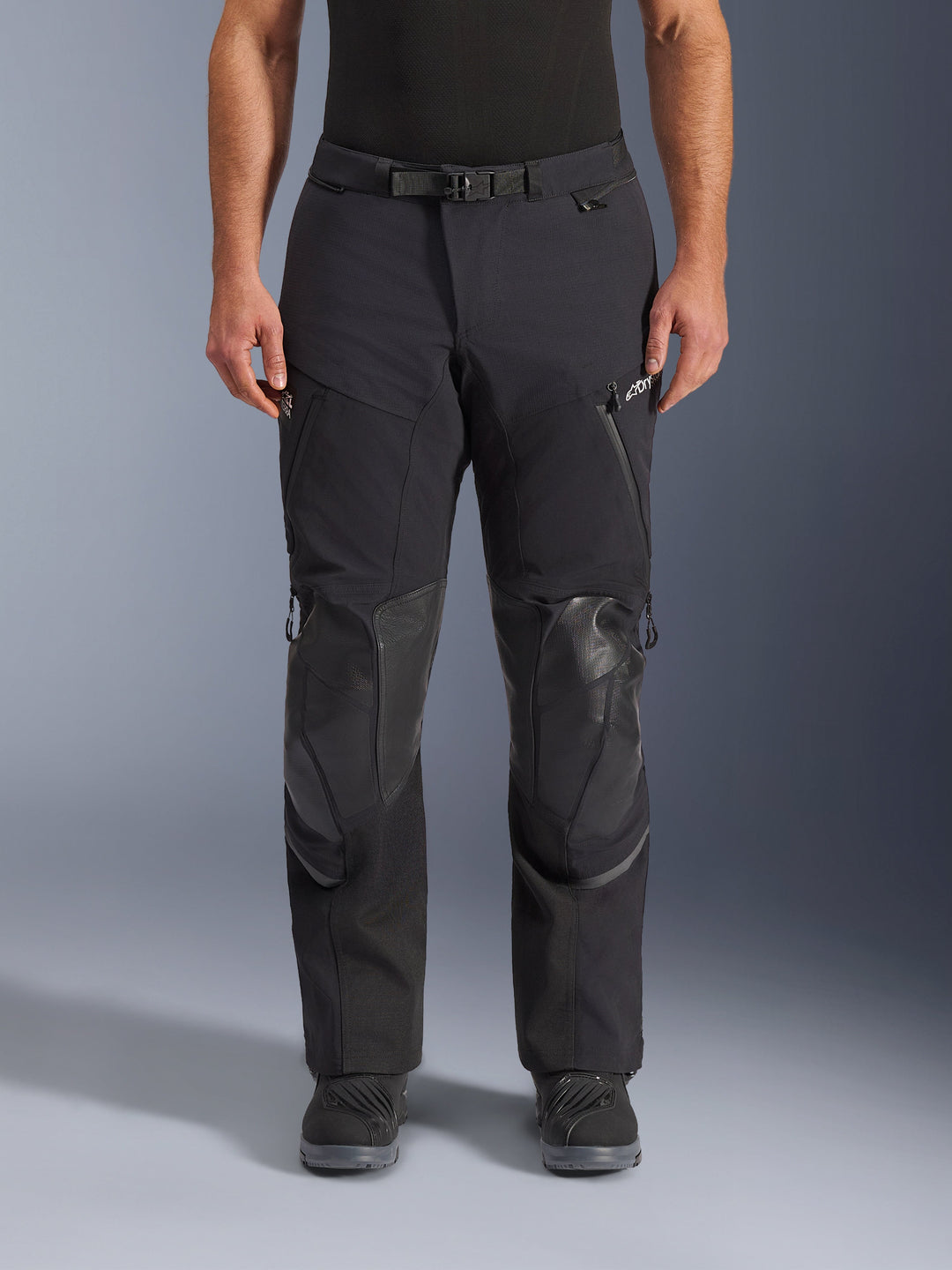 Pantalon Alpinestars Andes Pro Drystar® XF, pantalon de moto adventure touring noir et gris foncé, vue de face, avec construction laminée 3 couches, protections de genoux intégrées, poches zippées imperméables et boucle de taille magnétique, porté avec des bottes de moto noires