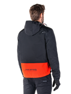 Alpinestars Mootant Waterproof Parka, Spicy Orange and Black, rear view on a male model, features a hooded black upper and bright orange lower section with MO.ST.EQ. branding, urban contemporary motorcycle jacket worn with black pants and gloves