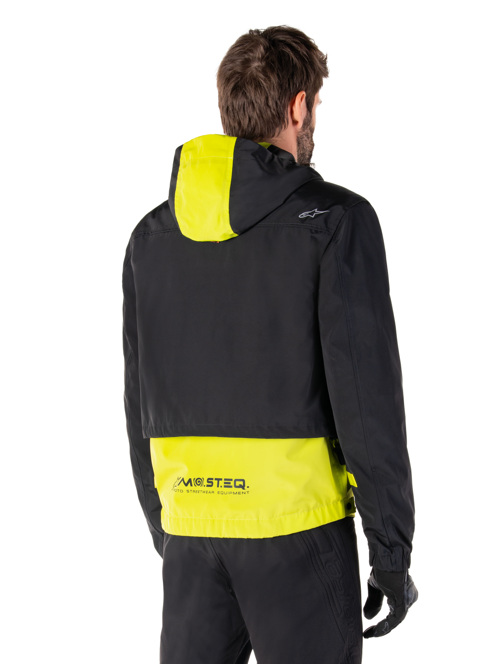 Parka imperméable Alpinestars Mootant, vue arrière, Lime Punch et noir, avec le haut du corps noir, un panneau de capuche et le bas du dos jaune citron, marquage MO.ST.EQ. sur l'ourlet, portée avec un pantalon de moto et des gants noirs, veste de moto urbaine contemporaine