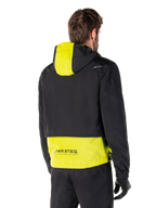 Parka imperméable Alpinestars Mootant, vue arrière, Lime Punch et noir, avec le haut du corps noir, un panneau de capuche et le bas du dos jaune citron, marquage MO.ST.EQ. sur l'ourlet, portée avec un pantalon de moto et des gants noirs, veste de moto urbaine contemporaine