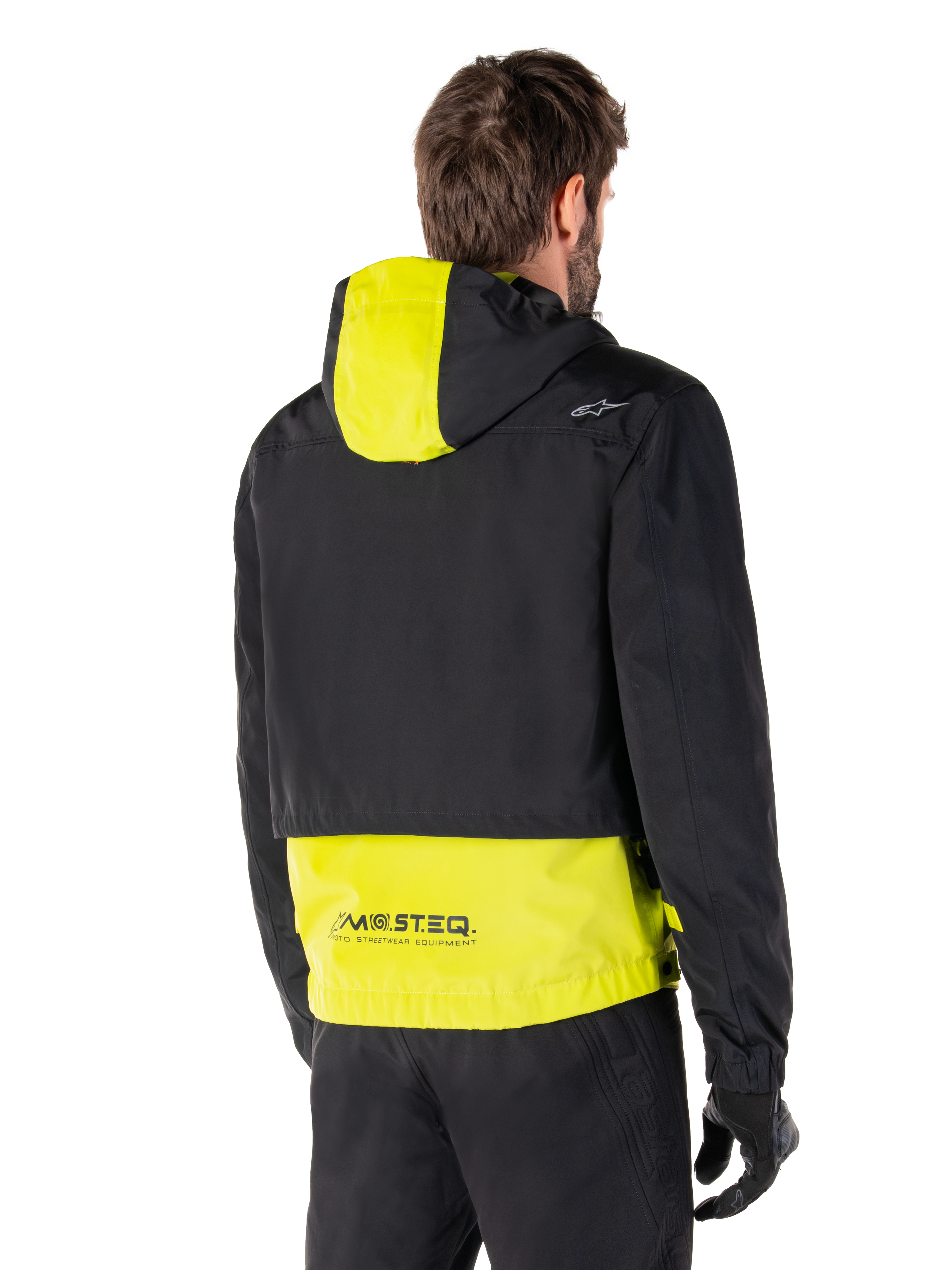 Parka imperméable Alpinestars Mootant, vue arrière, Lime Punch et noir, avec le haut du corps noir, un panneau de capuche et le bas du dos jaune citron, marquage MO.ST.EQ. sur l'ourlet, portée avec un pantalon de moto et des gants noirs, veste de moto urbaine contemporaine
