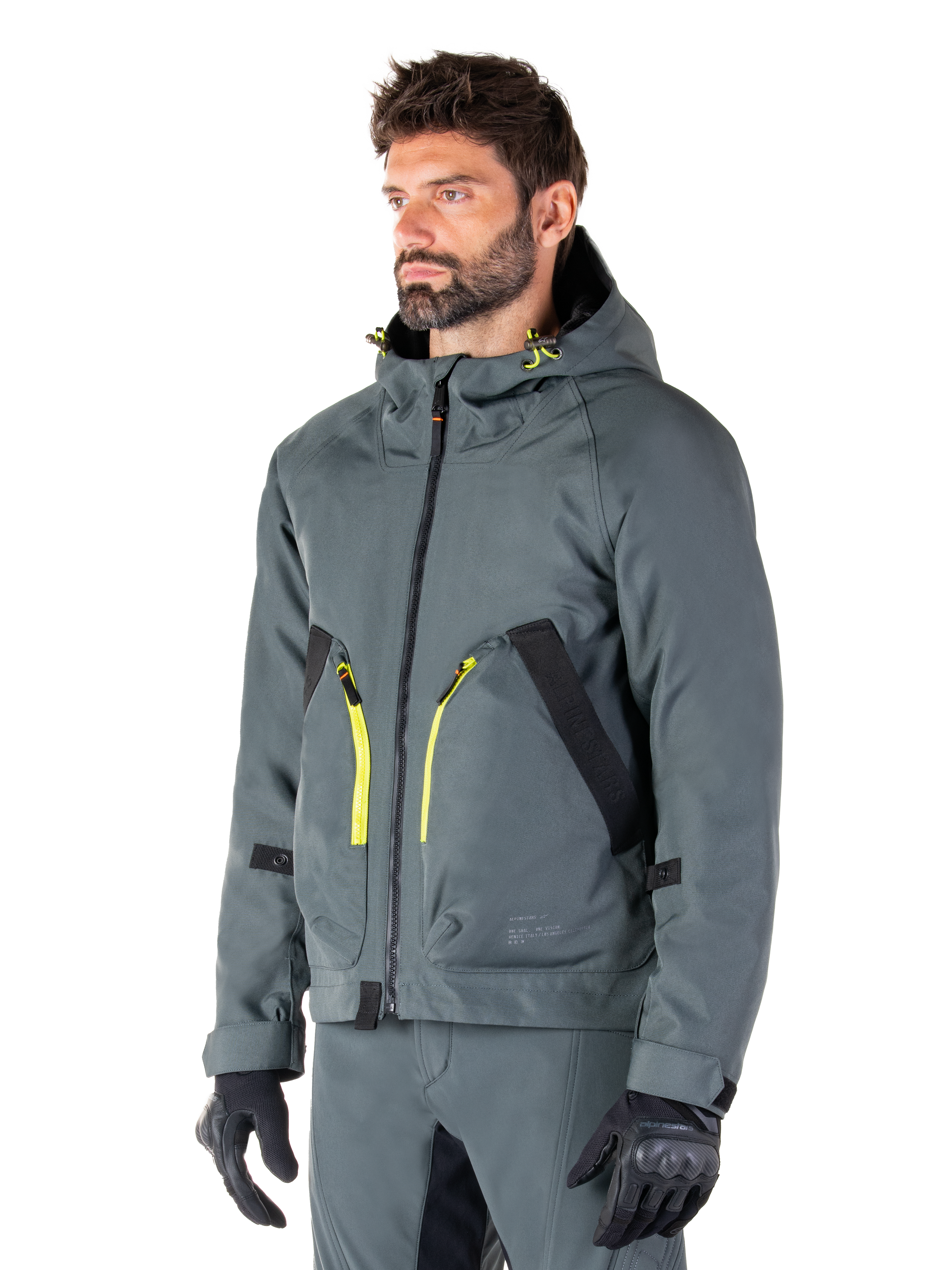 Parka imperméable Alpinestars Morush, veste de moto Urban Gray, design à capuche avec accents de fermetures éclair jaune vif, détails de poches noirs, portée avec un pantalon de moto gris assorti et des gants noirs, vêtement de moto contemporain inspiré du streetwear.