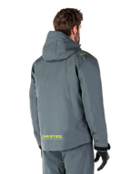 Parka imperméable Alpinestars Morush en Urban Gray, vue de dos, avec marquage MO.ST.EQ. jaune sur le bas du dos et l'épaule, design minimaliste à capuche, portée avec un pantalon de moto gris assorti et des gants noirs, style streetwear moto urbain