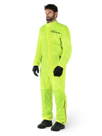 Combinaison Alpinestars Hurricane Rain V2, combinaison de moto imperméable une pièce, jaune fluo, portée avec des gants et des bottes noirs, avec un logo Alpinestars noir sur la poitrine et des détails réfléchissants sur les jambes