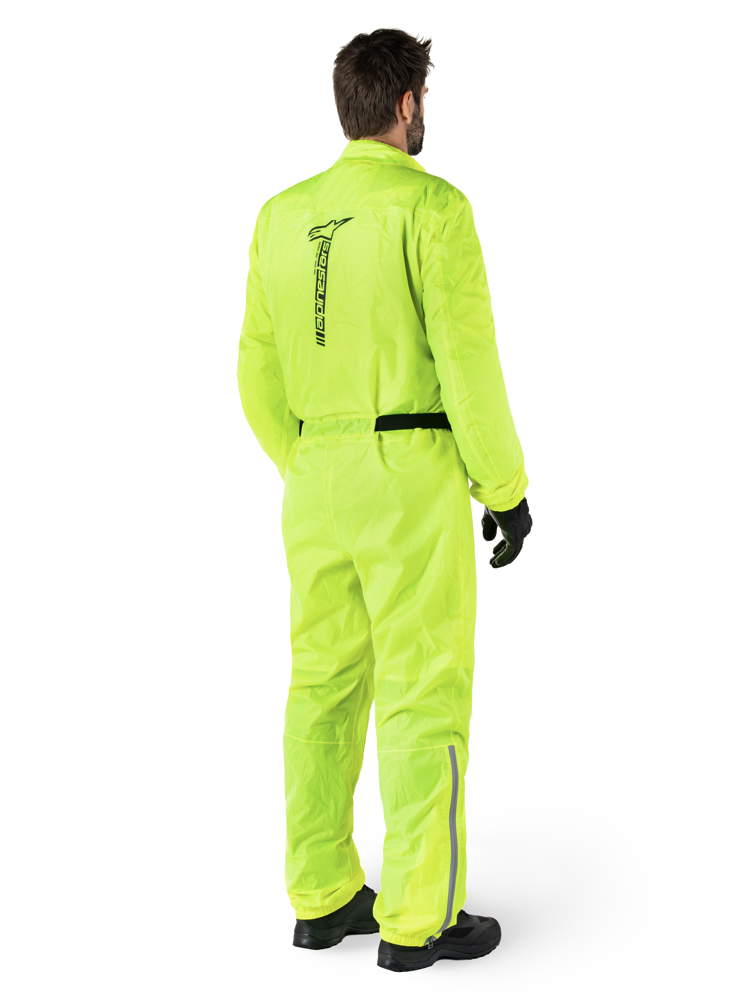 Combinaison Alpinestars Hurricane Rain V2, Jaune Fluo, vue arrière d'un motard portant une combinaison de pluie de moto une pièce, avec un logo Alpinestars noir au dos, une ceinture noire et des bandes réfléchissantes sur le bas des jambes, portée avec des gants et des bottes noirs