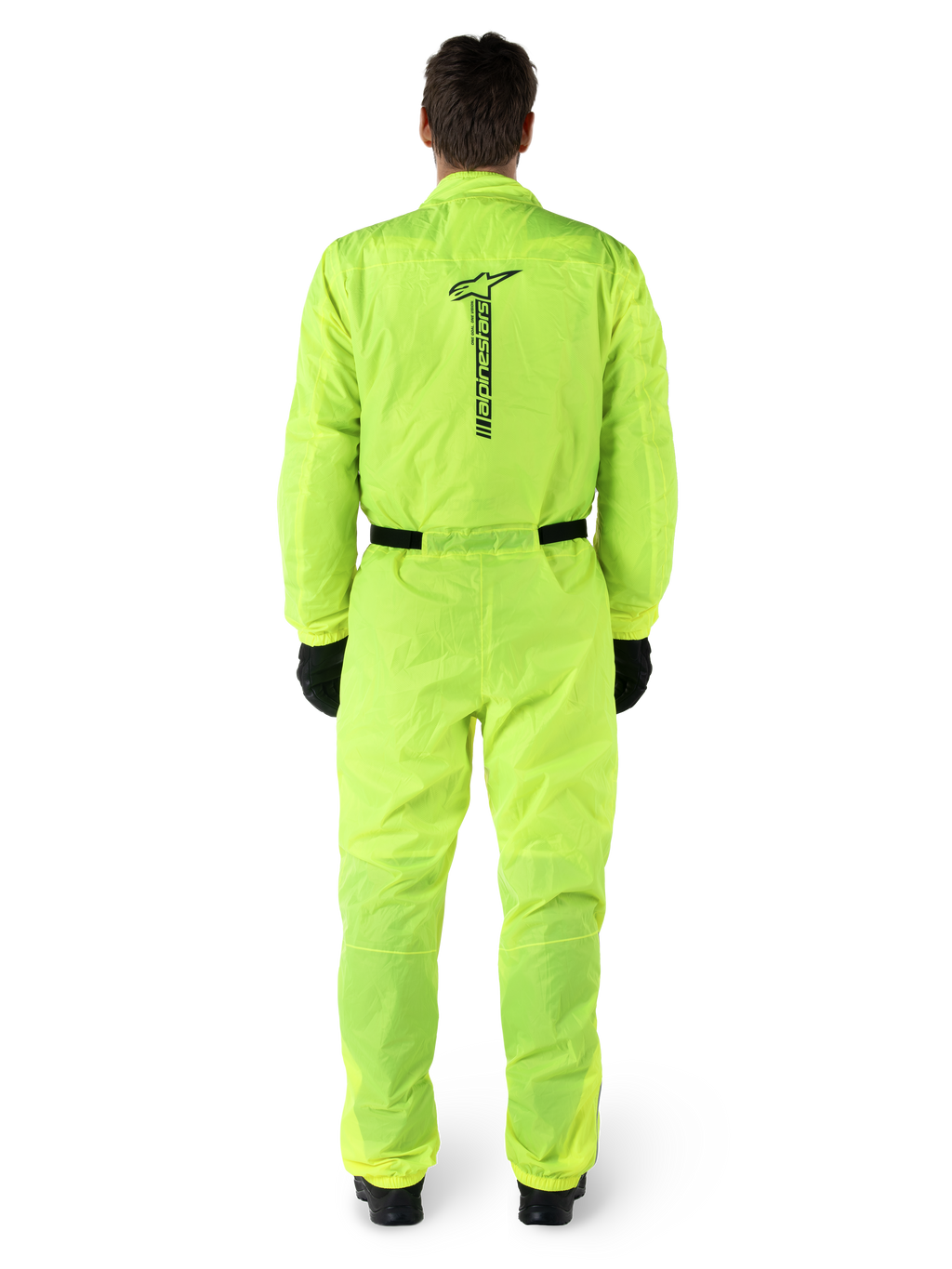Alpinestars Hurricane Rain V2 Suit, combinaison de pluie de moto une pièce, jaune fluo, vue arrière avec marquage Alpinestars noir vertical, taille et poignets élastiqués, portée avec des gants et des bottes noirs, équipement de conduite imperméable et coupe-vent