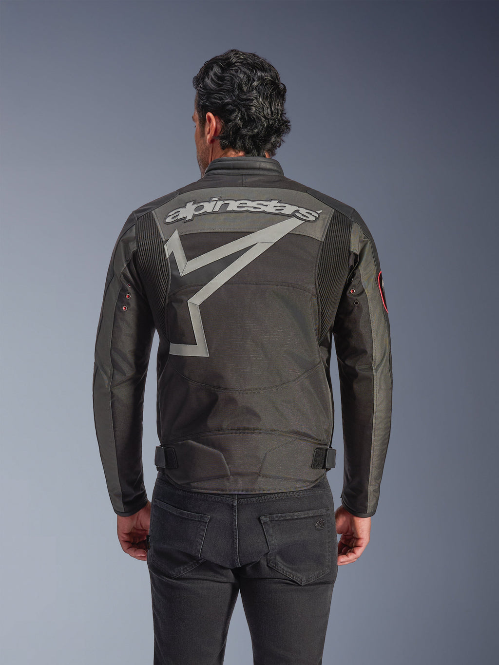 Veste Alpinestars Axton, veste de moto pour homme, Noir Anthracite Rouge Tangerine, vue arrière présentant un grand marquage Alpinestars gris et un motif d'étoile stylisé, panneaux en accordéon aux épaules et œillets métalliques d'inspiration vintage sur les manches.