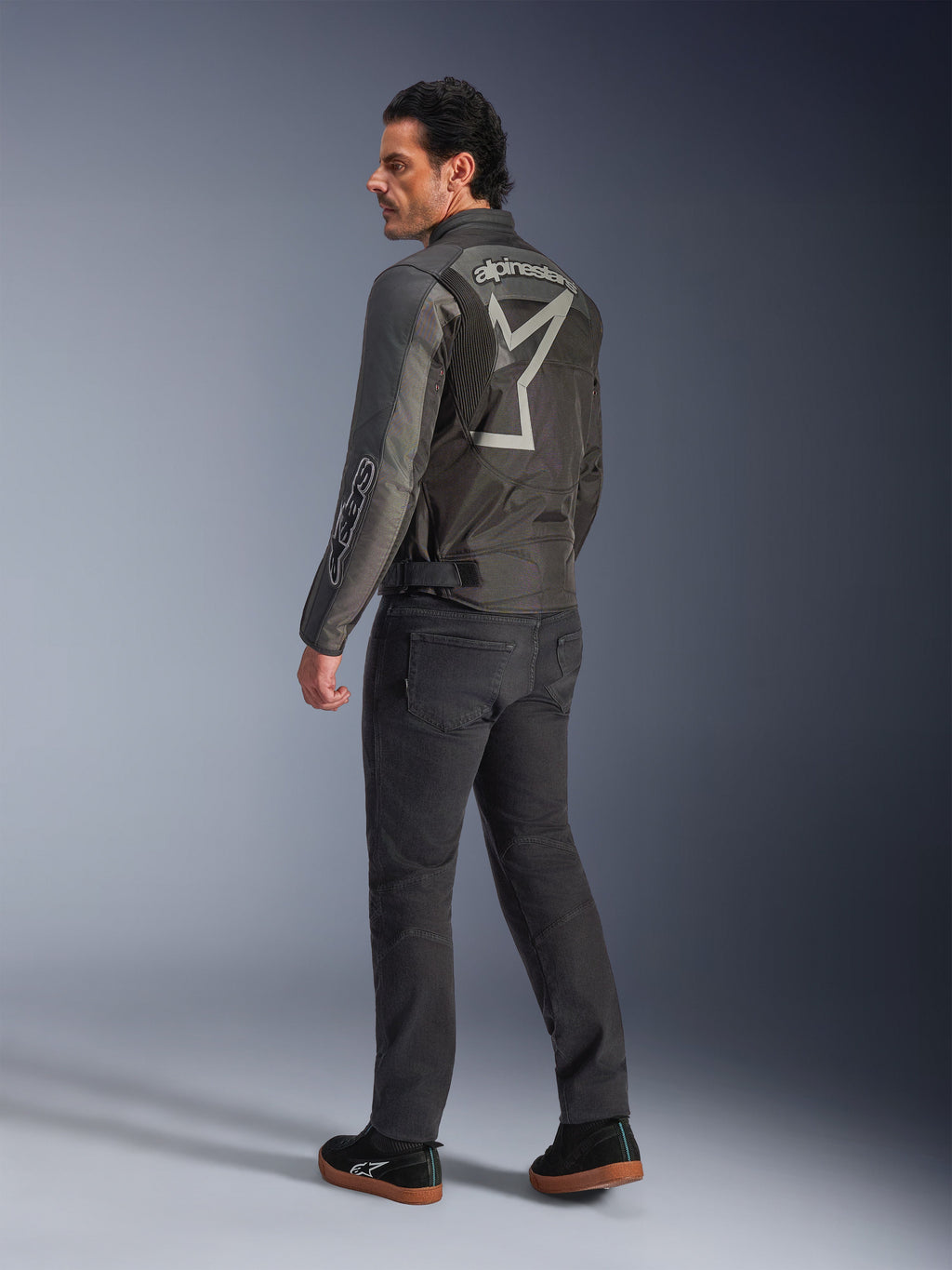 Veste Alpinestars Axton, veste de moto pour homme, noir et anthracite, vue arrière présentant un grand logo gris enveloppant et des panneaux accordéon aux épaules, portée avec un pantalon de moto sombre et des baskets noires, style urbain contemporain avec un héritage de course.