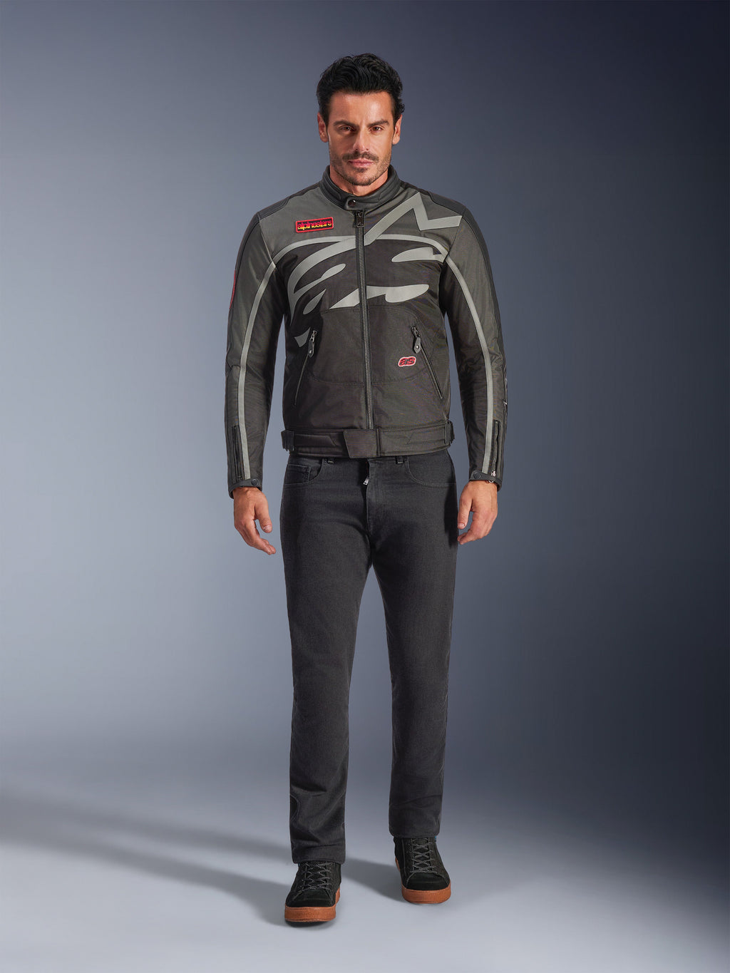 Veste Alpinestars Axton, veste de moto, Noir Anthracite Rouge Tangerine, portée par un mannequin avec un pantalon sombre, dotée d'un logo Alpinestars gris enveloppant, de patchs rouges de la marque et d'une coque en textile résistante à l'eau pour la conduite urbaine