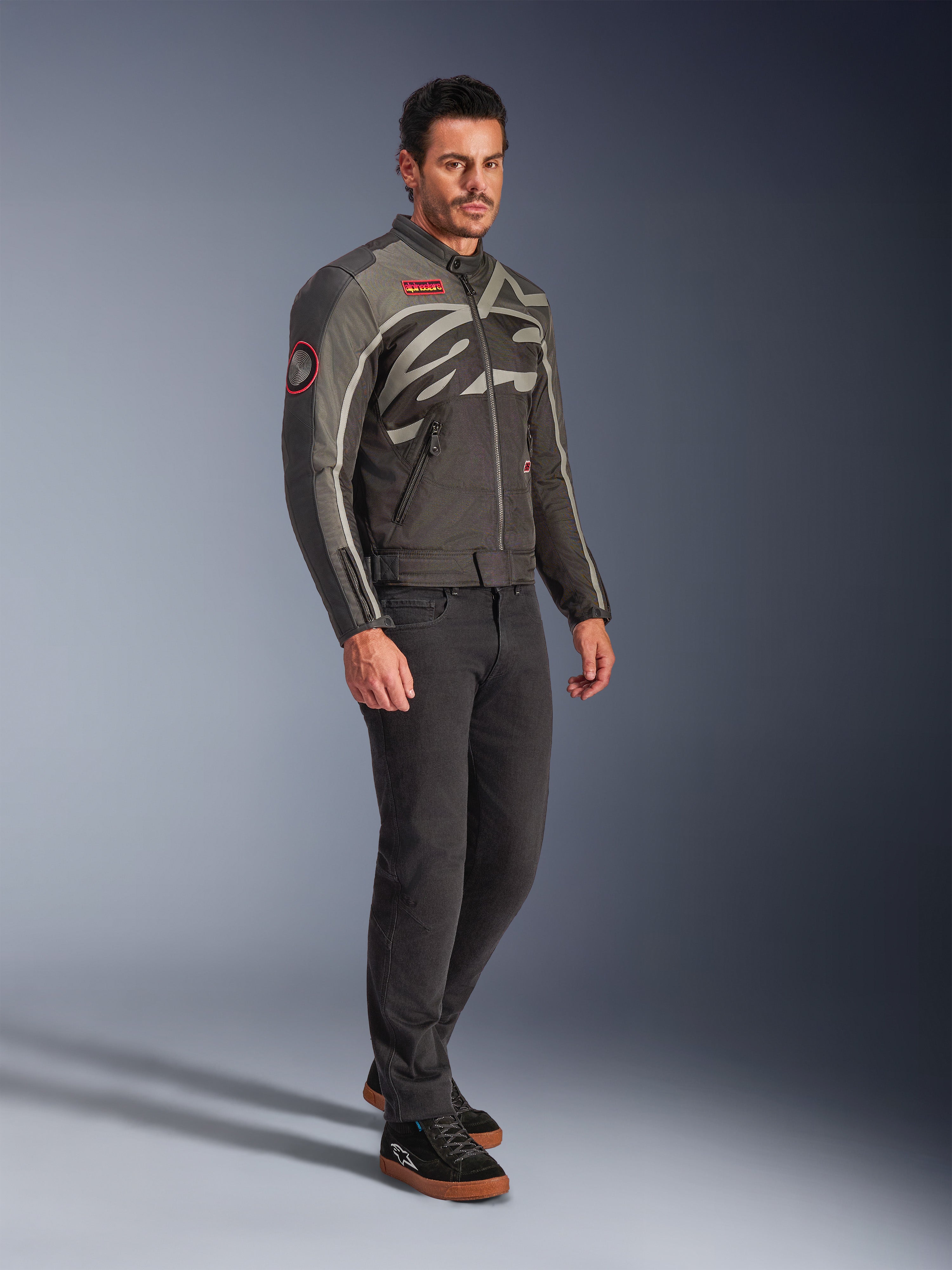 Modèle masculin portant la veste Alpinestars Axton en Noir Anthracite Rouge Tangerine, veste de moto d'inspiration vintage avec un logo enveloppant et des accents rouges, associée à un pantalon noir et des baskets de moto noires, équipement de moto urbain contemporain
