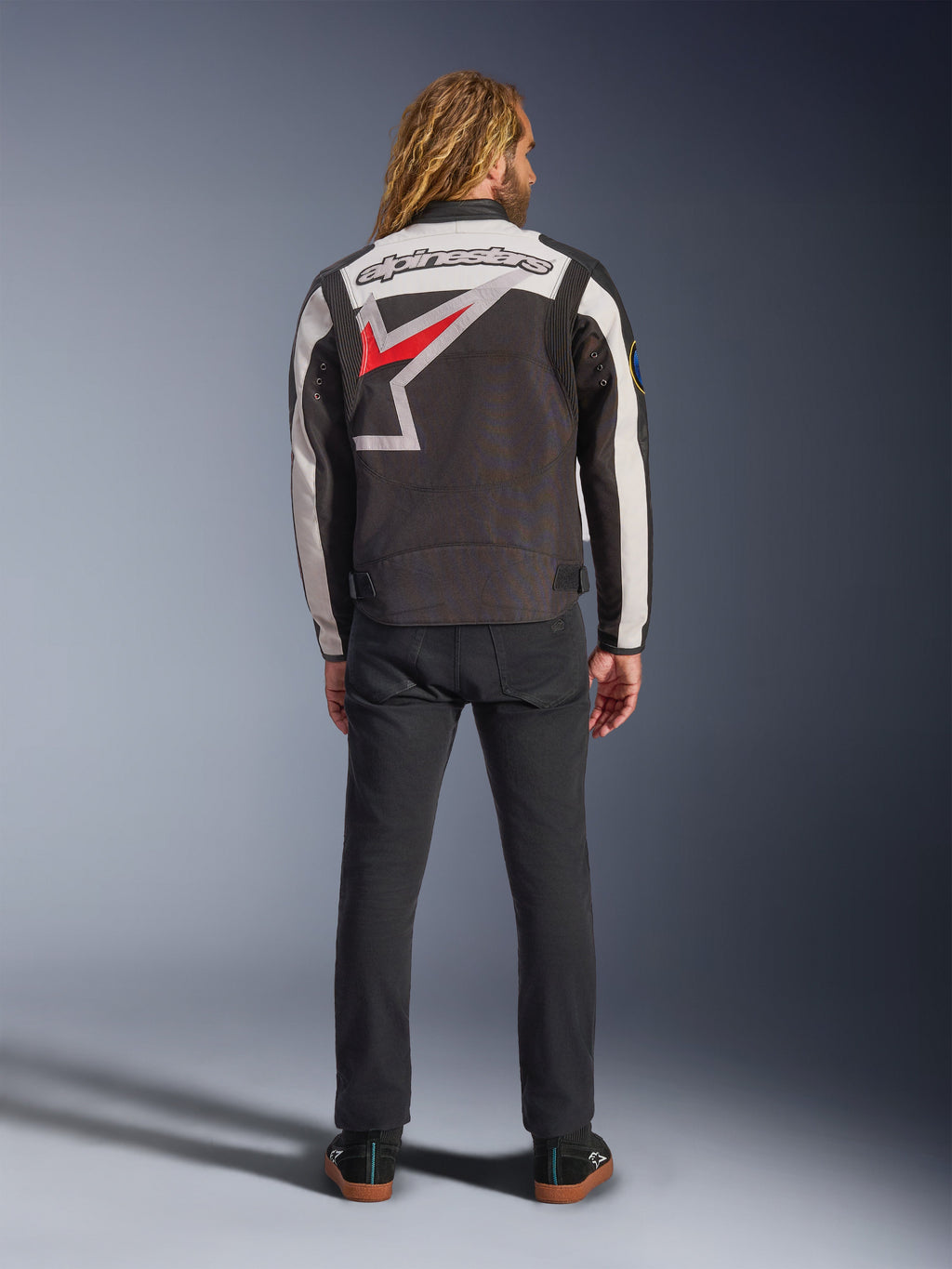 Veste Alpinestars Axton, veste de moto, noir, blanc cassé et rouge vif, vue de dos sur un mannequin, avec un grand logo étoile Alpinestars dans le dos, empiècements de manches blanc cassé, portée avec un pantalon noir, design inspiré des vêtements d'équipe de course vintage