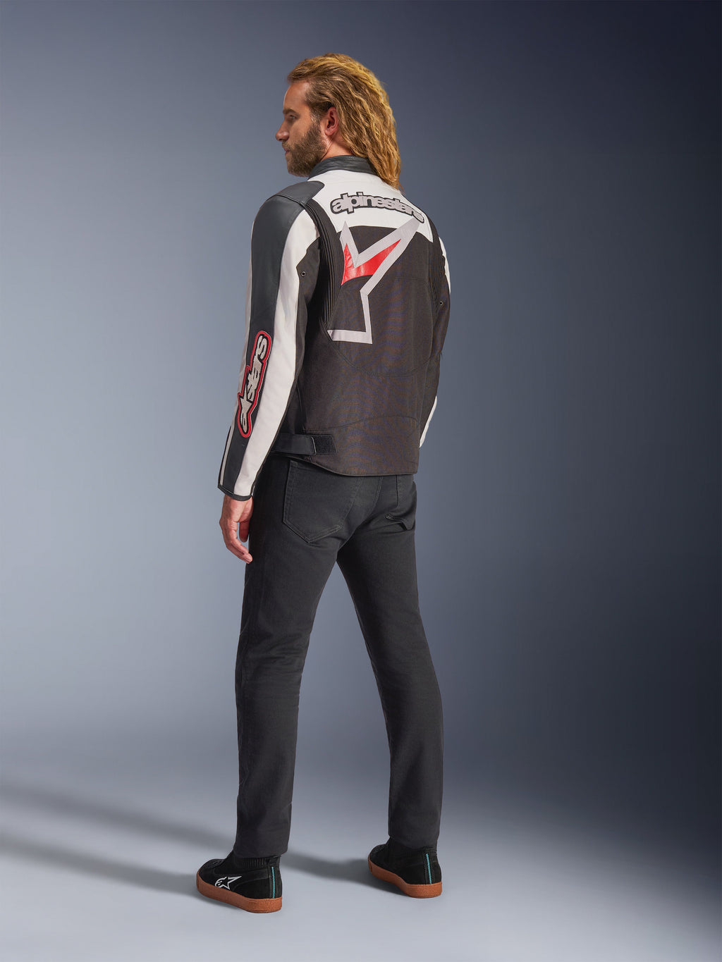 Veste Alpinestars Axton, veste de moto, noir, blanc cassé et rouge vif, vue arrière portée par un mannequin homme, arborant un grand logo étoile et le nom de la marque Alpinestars dans le dos, design d'inspiration vintage racing avec des panneaux en accordéon, associée à un pantalon noir et des baskets