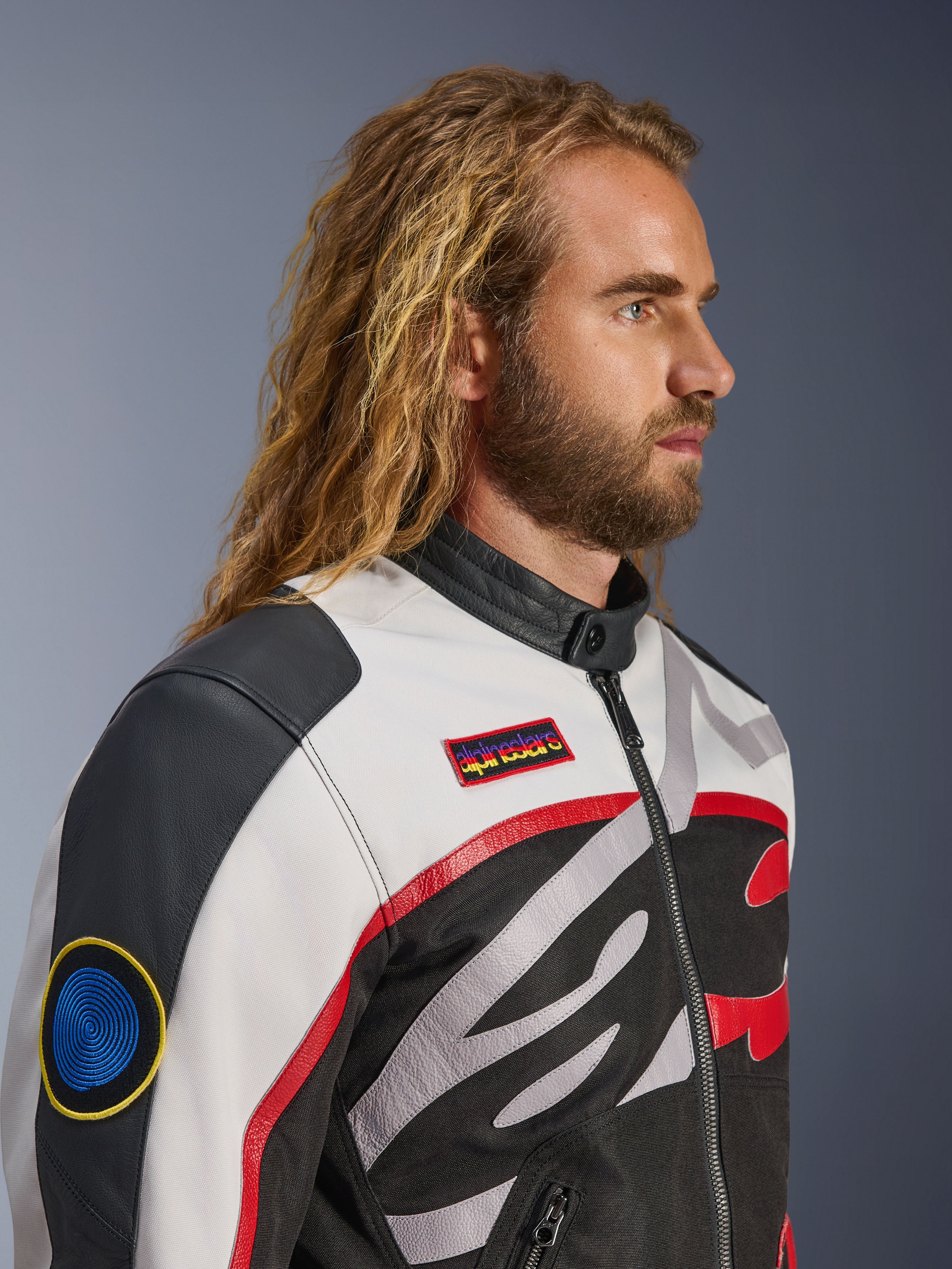 Homme aux cheveux longs et barbu portant la veste Alpinestars Axton, veste de moto, noir, blanc cassé et rouge vif, design d'inspiration racing vintage avec écussons de la marque et logo enveloppant, vue de profil rapprochée, style urbain contemporain