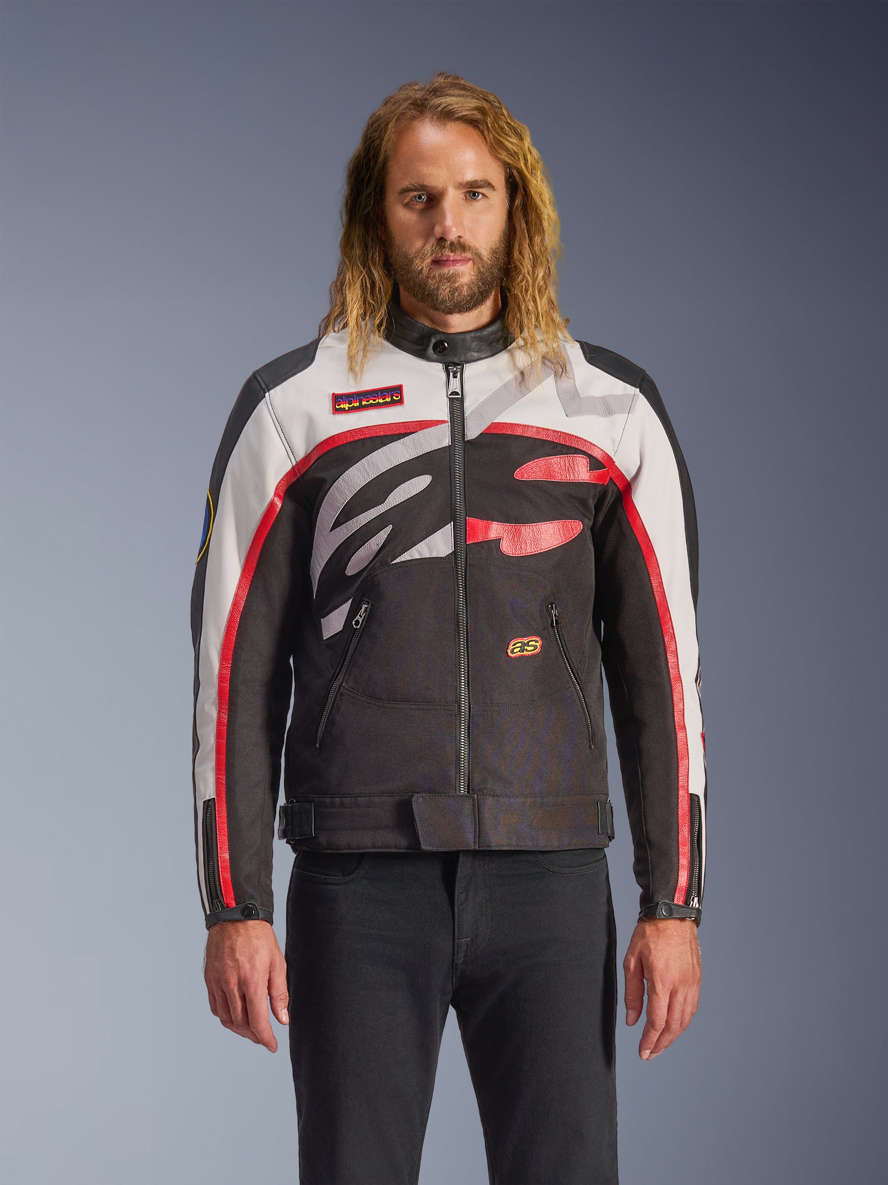 Alpinestars Axton Jacket, veste de moto, noir, blanc cassé et rouge vif, avec des motifs abstraits audacieux sur la poitrine et un logo enveloppant, manches à rayures rouges, design d'inspiration vintage avec un col minimaliste, portée par un mannequin avec un jean noir.