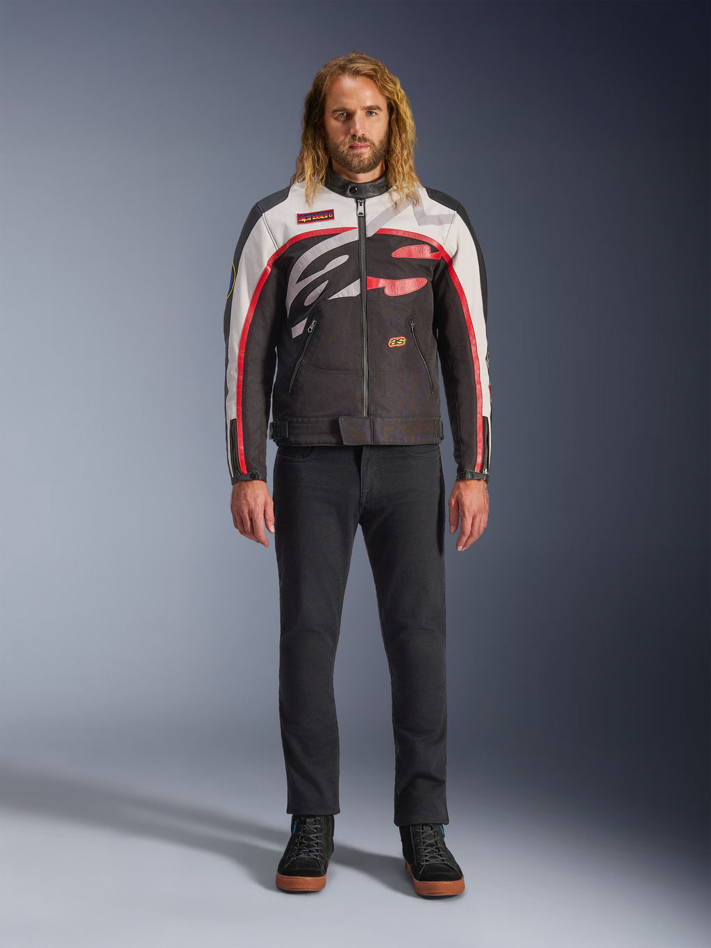 Veste Alpinestars Axton, veste de moto, noir, blanc cassé et rouge vif, portée par un mannequin avec un pantalon noir et des baskets, dotée d'un logo enveloppant audacieux et d'un design inspiré de la course vintage