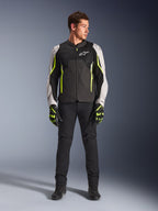 Modèle masculin portant la veste Alpinestars AST V3 Air en Noir Argent Jaune Fluo, dotée de panneaux de ventilation en mesh et d'accents fluorescents, associée à un pantalon de moto noir et des gants assortis, équipement de performance pour conduite sportive.