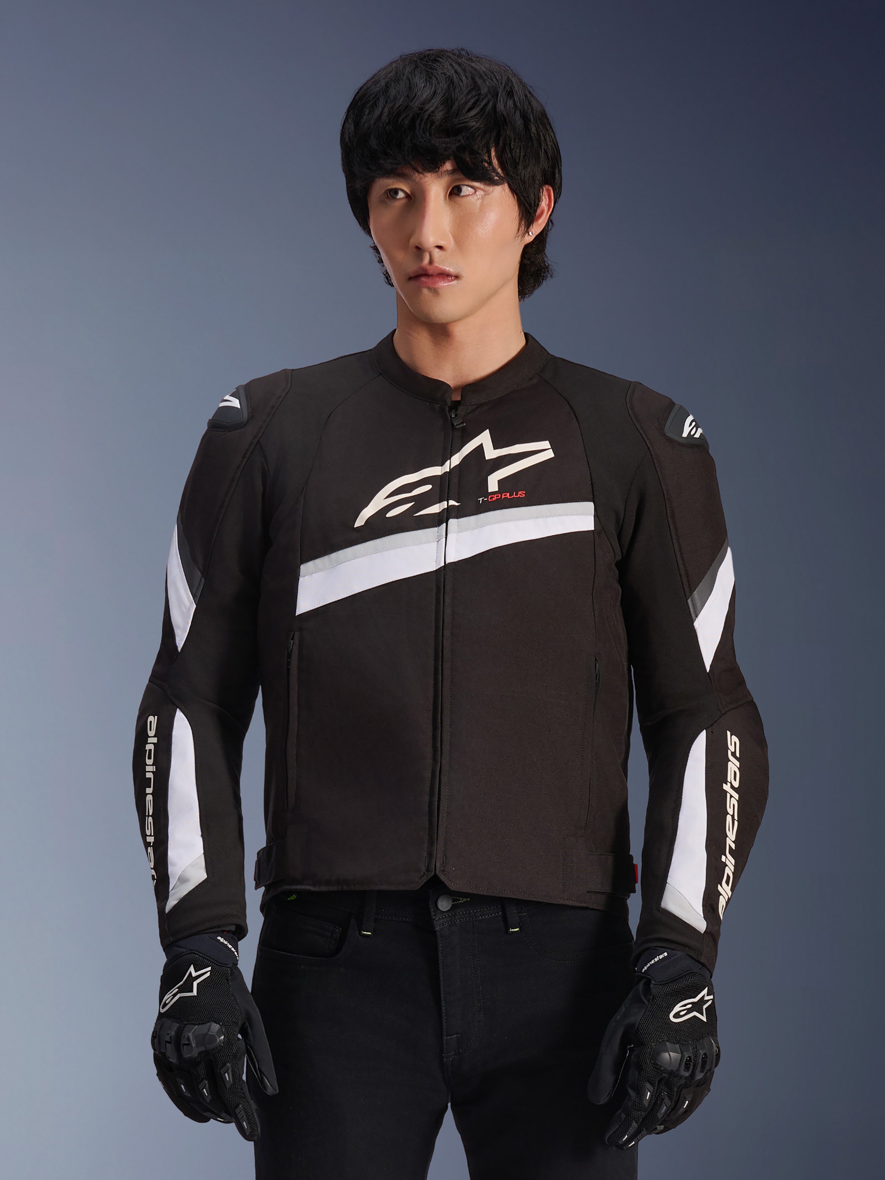Veste Alpinestars T-GP Plus R V4, noir et blanc, veste de moto en textile avec accents diagonaux blancs et logos de la marque sur la poitrine et les manches, portée par un mannequin avec des gants de moto noirs, coupe sportive épurée pour la route