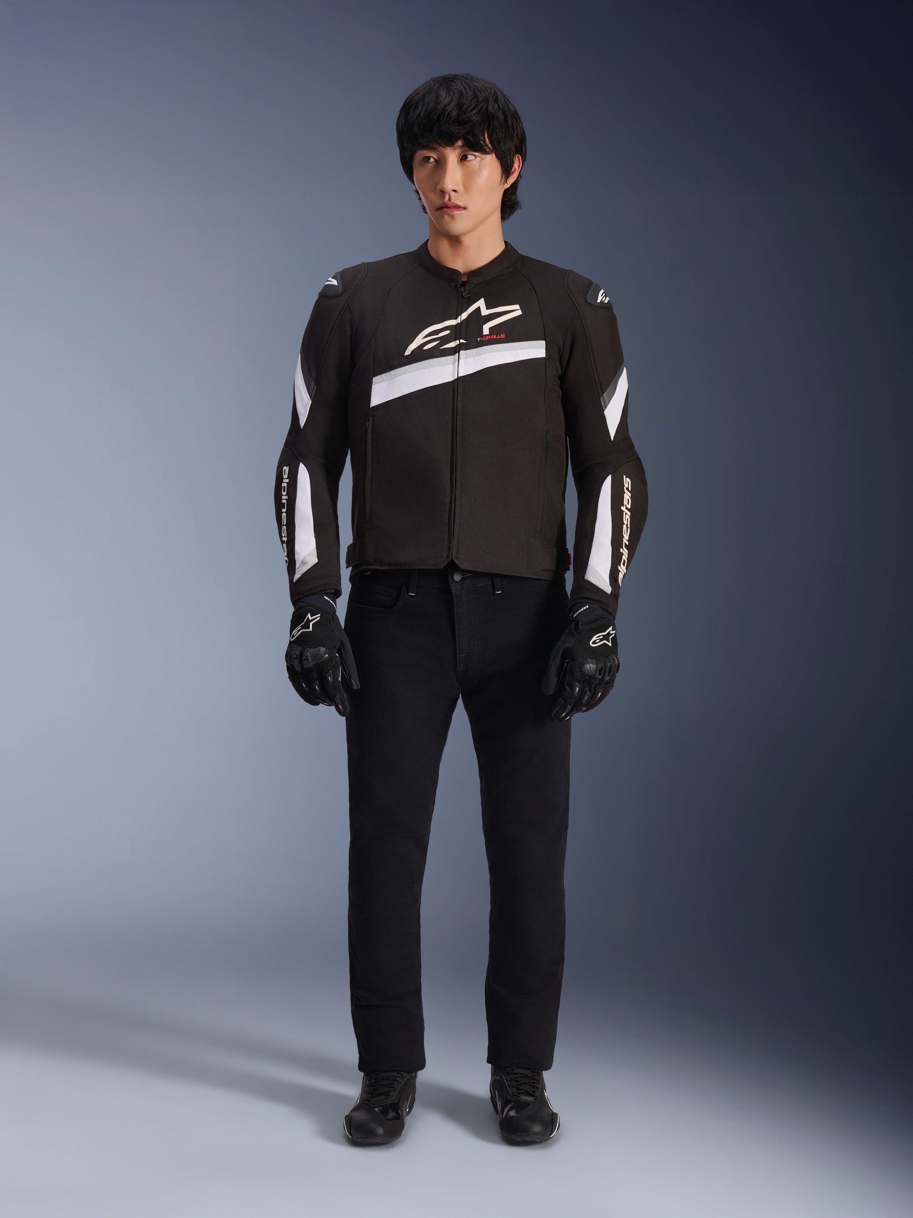 Veste Alpinestars T-GP Plus R V4, noir et blanc, veste de moto en textile sport, portée par un mannequin avec un pantalon et des gants noirs, logo Alpinestars blanc sur la poitrine et les manches, design épuré inspiré de la course