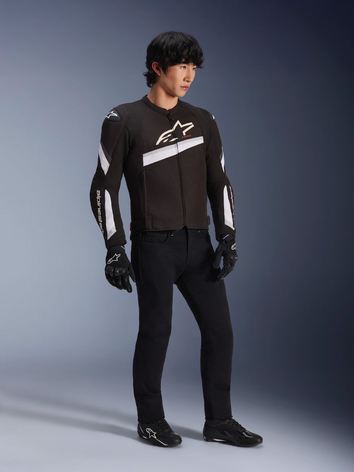 Veste Alpinestars T-GP Plus R V4, veste de moto en textile noir et blanc portée par un mannequin avec un pantalon de moto noir et des gants, avec des logos Alpinestars blancs sur la poitrine et les manches, design sportif pour la performance sur route