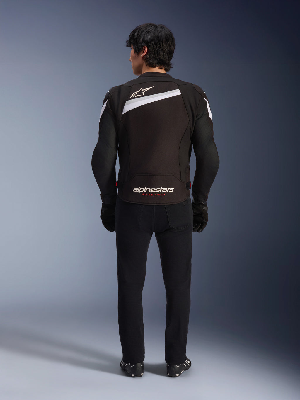 Veste Alpinestars T-GP Plus R V4, veste de moto sport en textile, noir et blanc, vue arrière avec logo Alpinestars blanc et marquage 'RACING AHEAD', portée avec un pantalon et des bottes noirs, design profilé pour la conduite de performance.