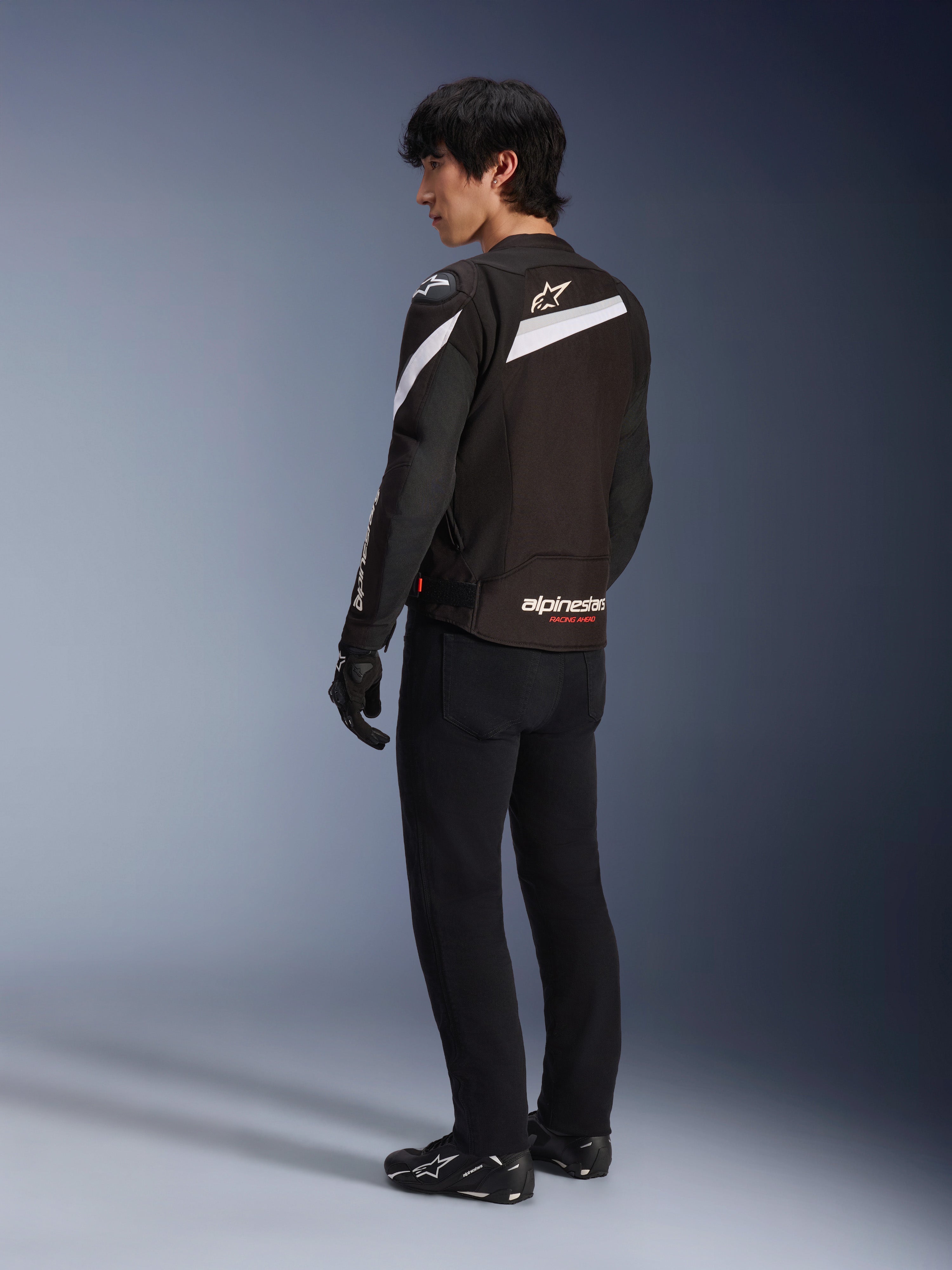 Pilote portant la veste Alpinestars T-GP Plus R V4 en Noir et Blanc, vue arrière montrant les accents diagonaux blancs et le logo sur le bas du dos, associée à un pantalon de moto noir, des gants et des bottes techniques, équipement de moto en textile coupe sport