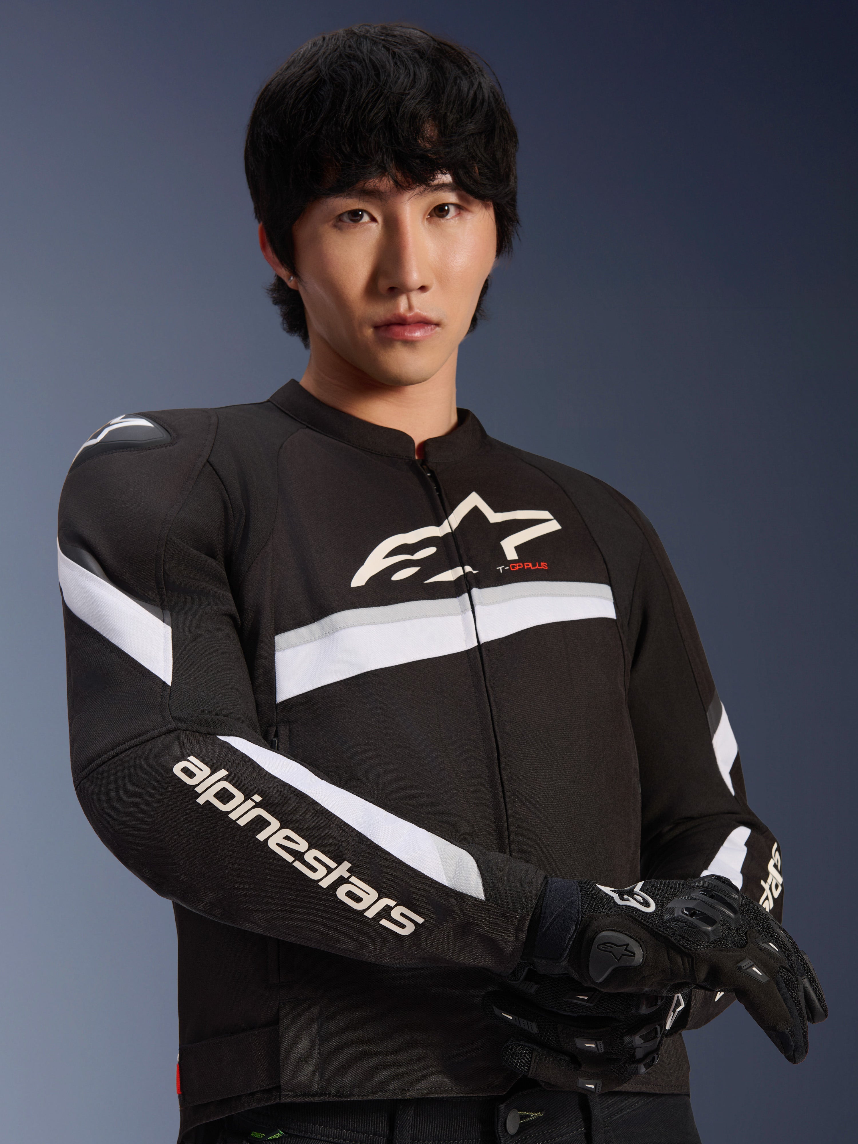 Veste Alpinestars T-GP Plus R V4, noir et blanc, portée par un mannequin homme, veste de moto en textile avec des rayures blanches sur la poitrine et les manches, logo Alpinestars sur la poitrine, associée à des gants de moto noirs