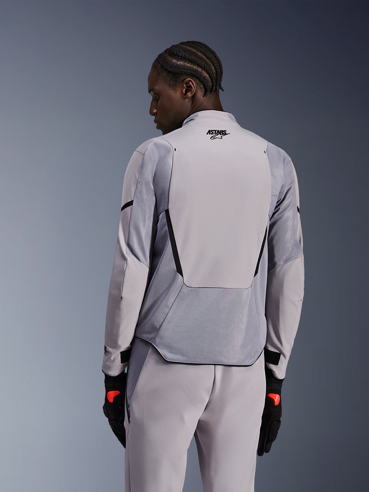 Veste Alpinestars Aeroshell Airflow en Gris Moyen Anthracite, vue de dos montrant les panneaux en maille respirante et les perforations au laser, construction en softshell technique extensible avec un panneau arrière allongé, portée avec un pantalon gris assorti et des gants noirs pour un look sportif.