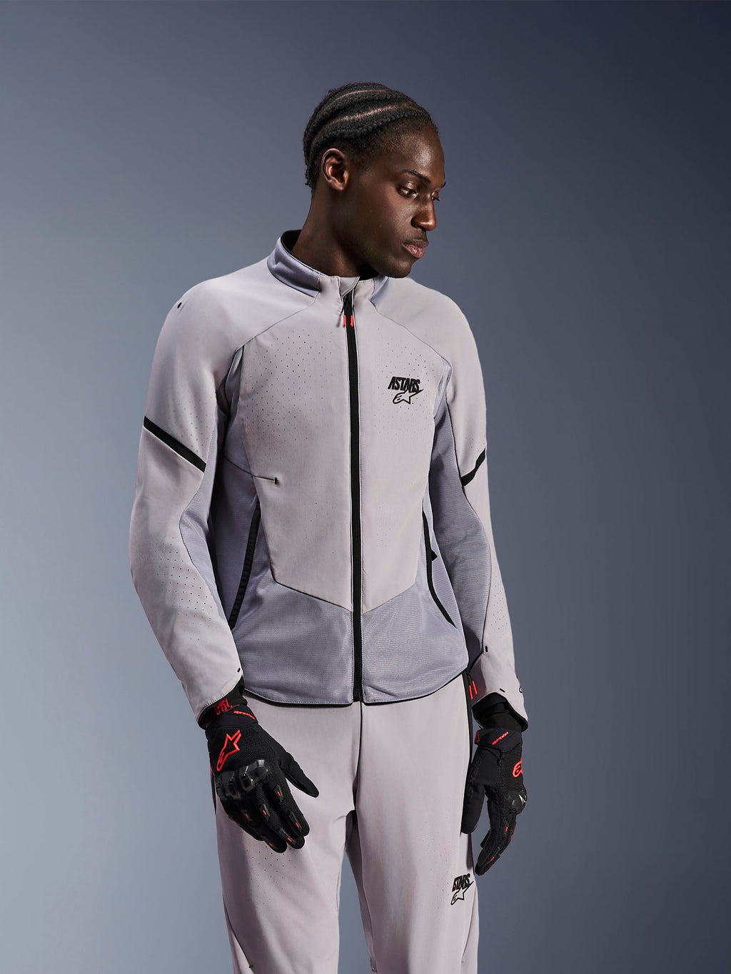 Veste Alpinestars Aeroshell Airflow, gris anthracite moyen, portée par un mannequin avec un pantalon textile assorti et des gants noirs avec des accents rouges, dotée de zones perforées au laser et de panneaux en maille pour la ventilation, design à coupe sportive pour la conduite moto.